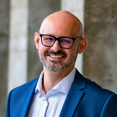 A smiling bald man with a beard, wearing glasses, a blue blazer, and a white shirt, standing in front of a stone wall.