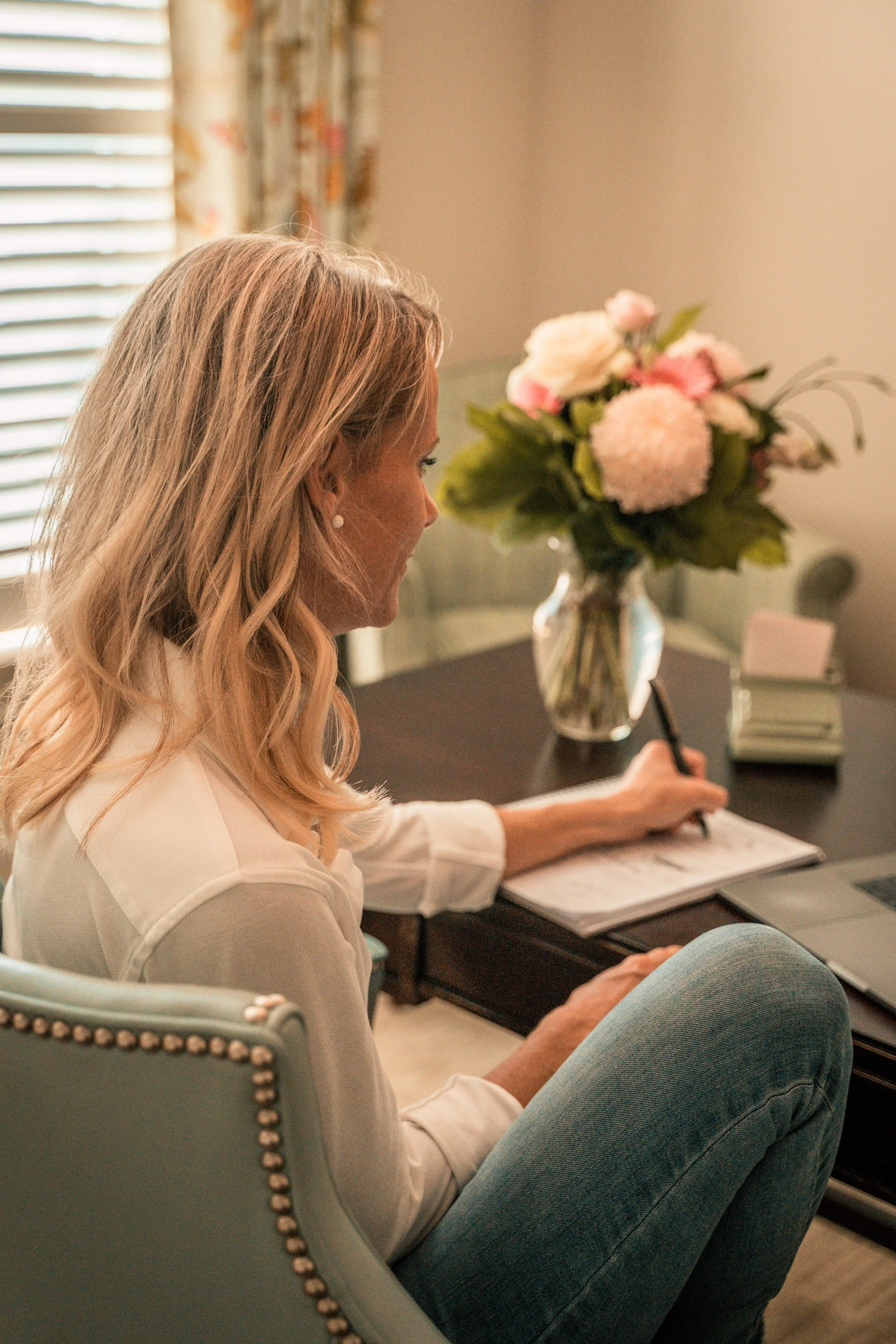 A woman with blonde hair, wearing a white shirt and light blue jeans, is sitting at a dark wooden table. She is writing in a notebook with a pen. A vase with pink and cream flowers is on the table, along with some books and a laptop. She appears to be in a well-lit room with a window, with floral curtains in the background.