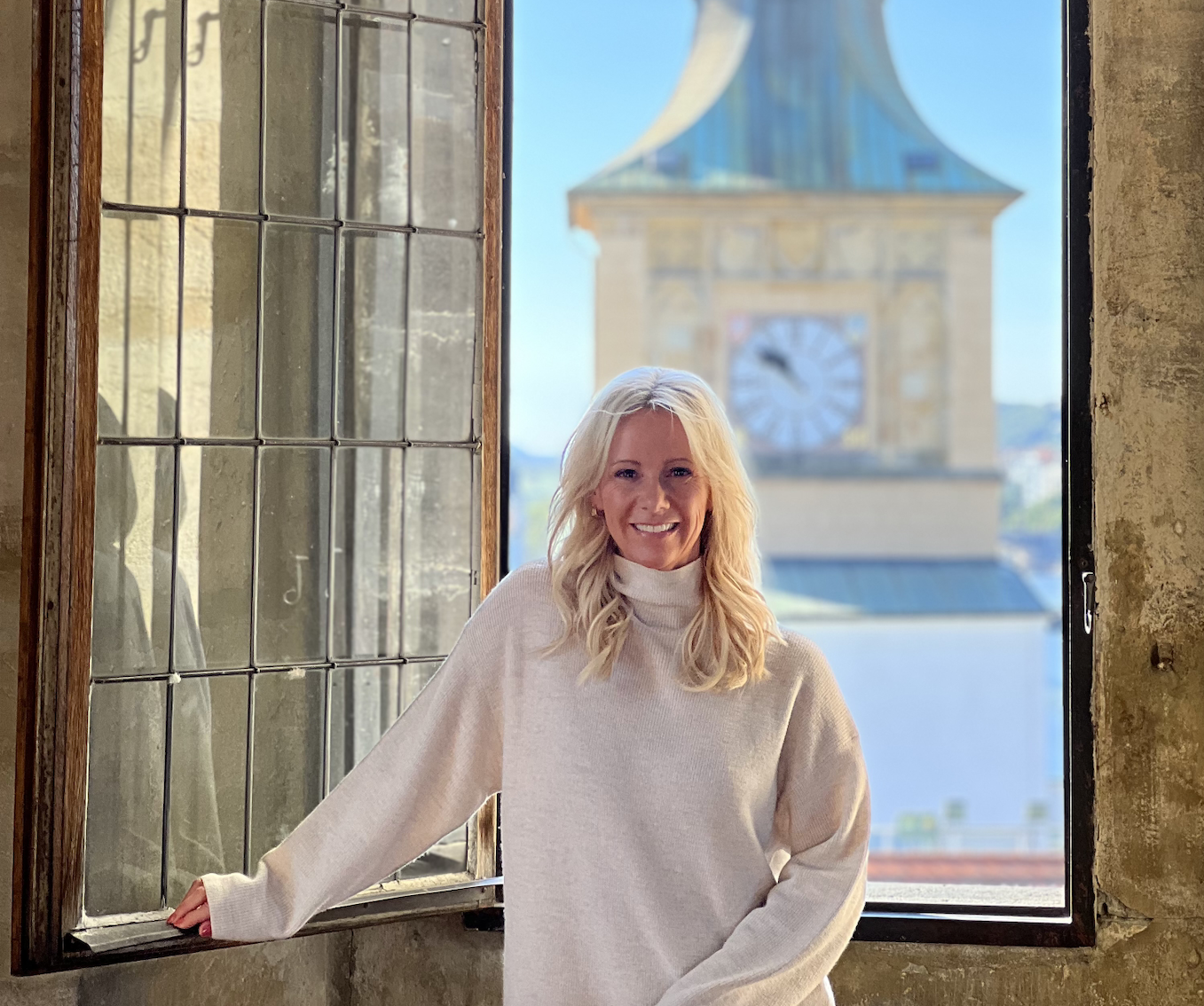 A smiling woman with blonde hair wearing a white turtleneck sweater, standing by an open window with a view of a clock tower in the background during daytime.