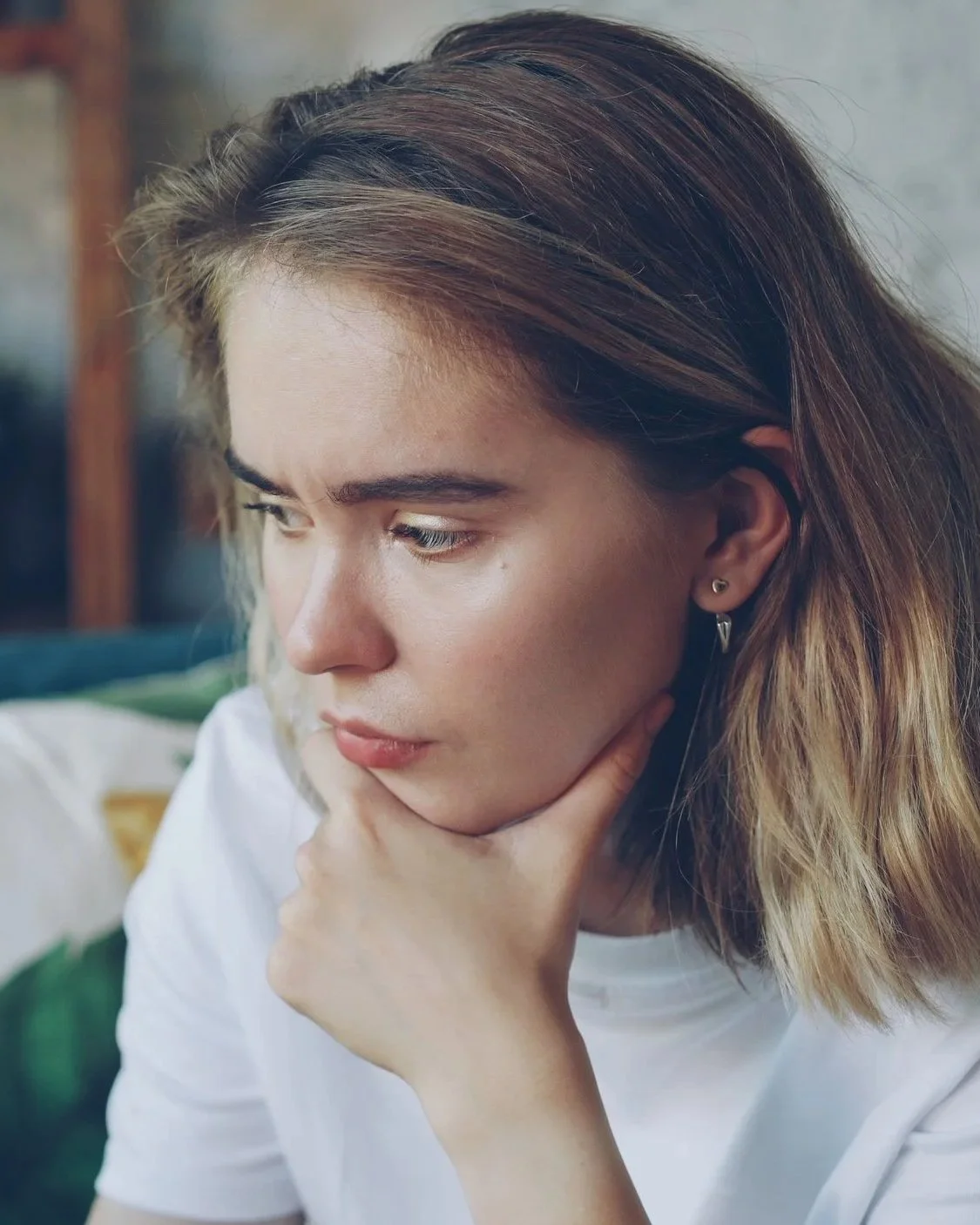 A young woman with brown hair and multiple earrings, resting her chin on her hand, looking thoughtful.