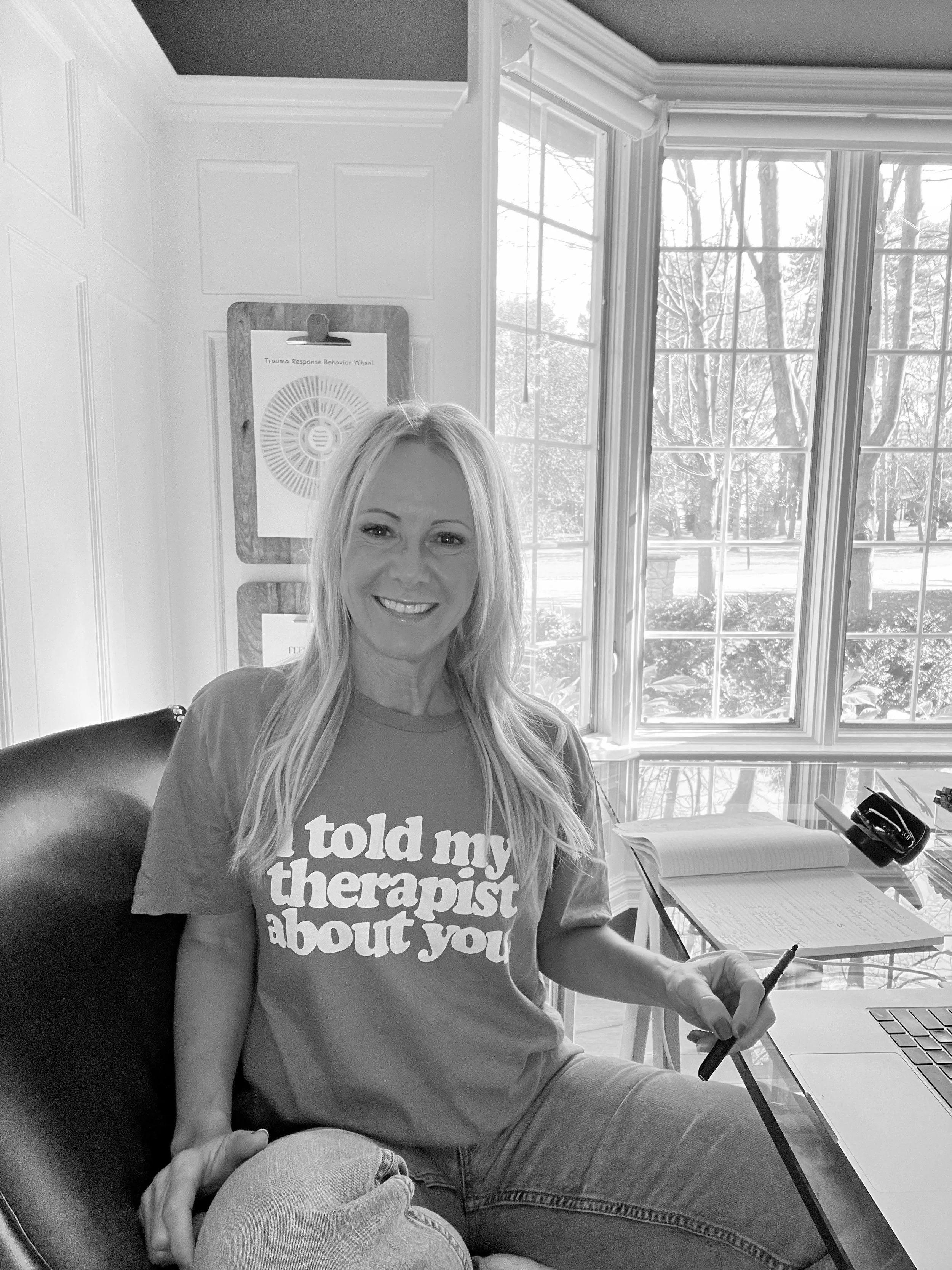 A woman smiling at a desk with sunlight coming through large windows behind her. She wears a t-shirt that says 'told my therapist about you' and holds a pen near a laptop. There are papers on the desk and a hand resting on her knee.
