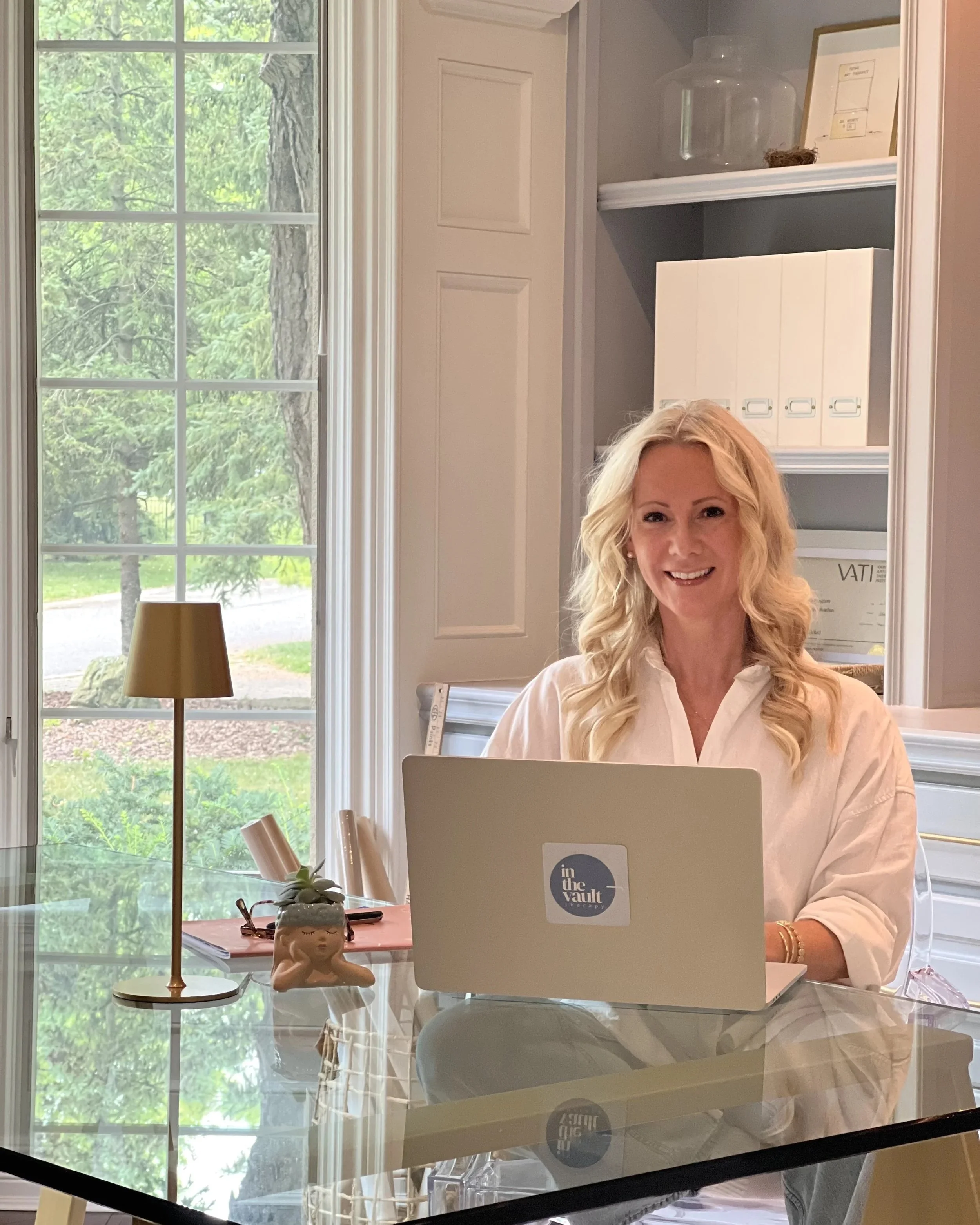 Woman with long blond curly hair smiling at a laptop on a glass table in a bright room with large windows showing trees outside. There is a small decorative figure on the table and a lamp nearby.