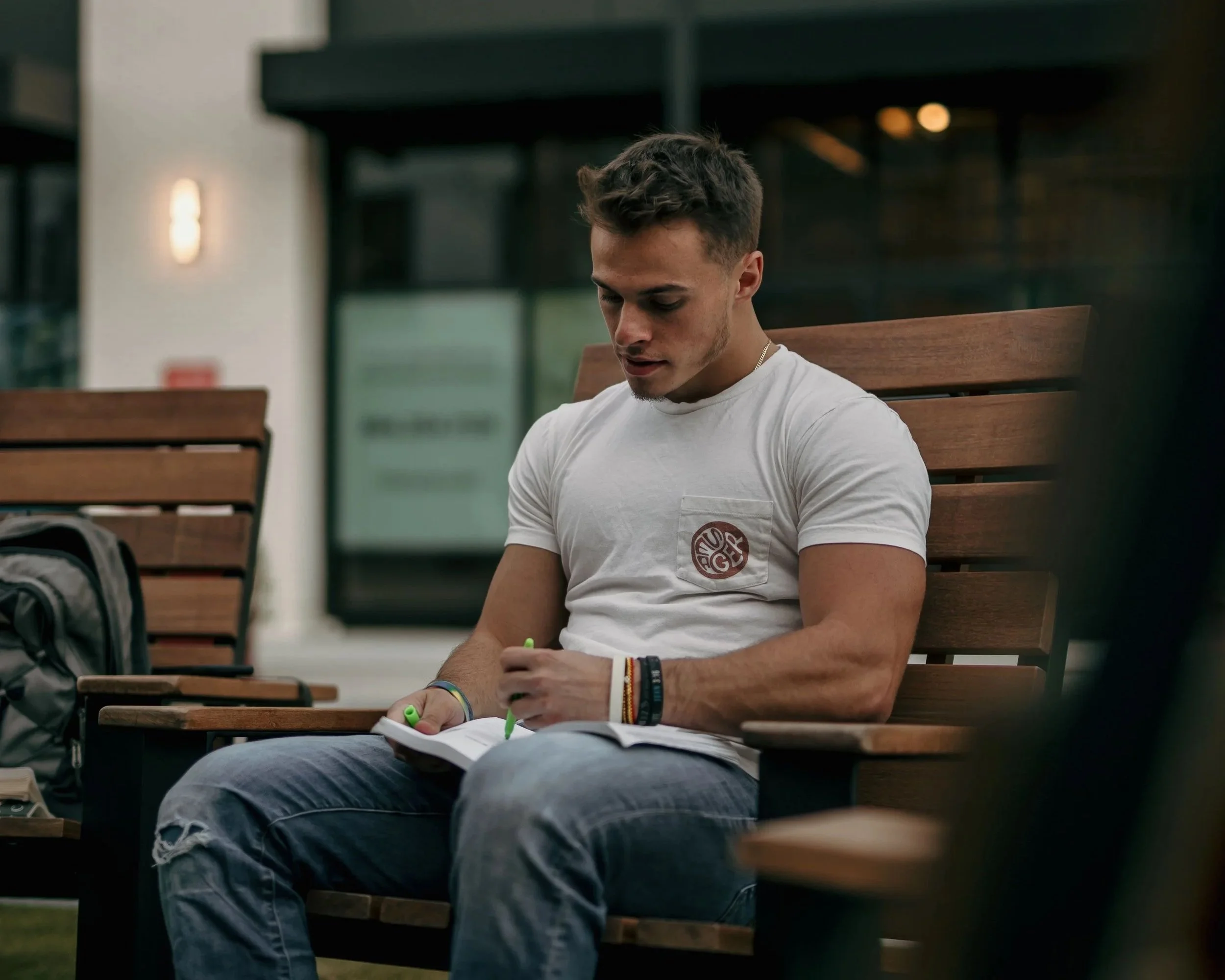 A young man with short brown hair, wearing a white t-shirt and jeans, sitting on a wooden bench outside, reflecting upon life in a notebook.