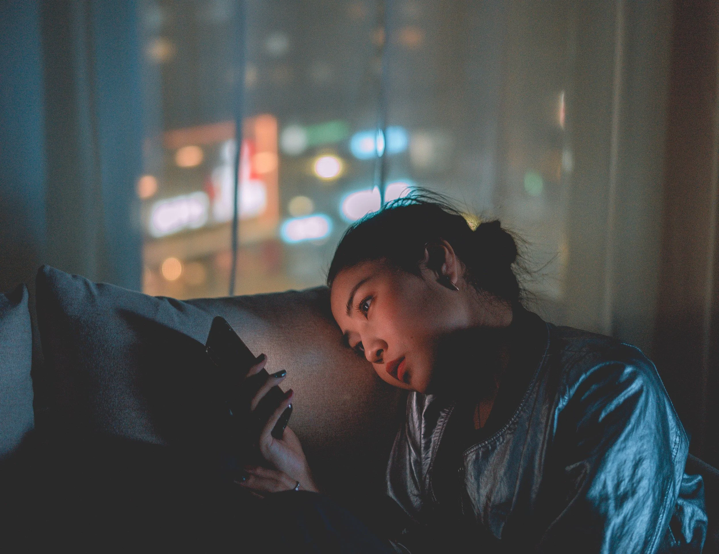 A woman with dark hair is lying on her side in a dimly lit room, staring at her phone which she holds in her hand. The background shows colorful blurred lights through a window.