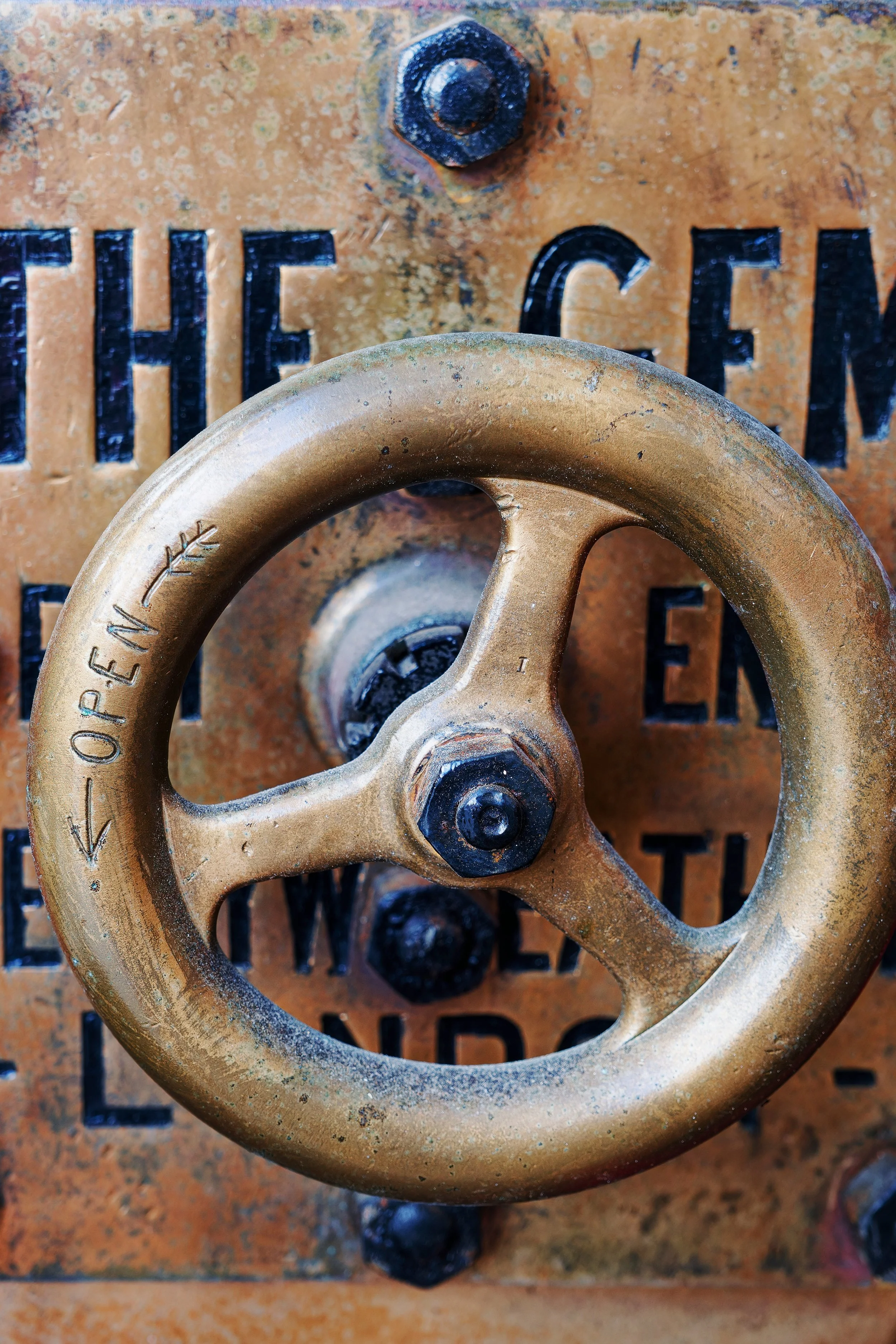 Close-up of a weathered brass valve wheel attached to a rusted metal surface with partially visible black text.