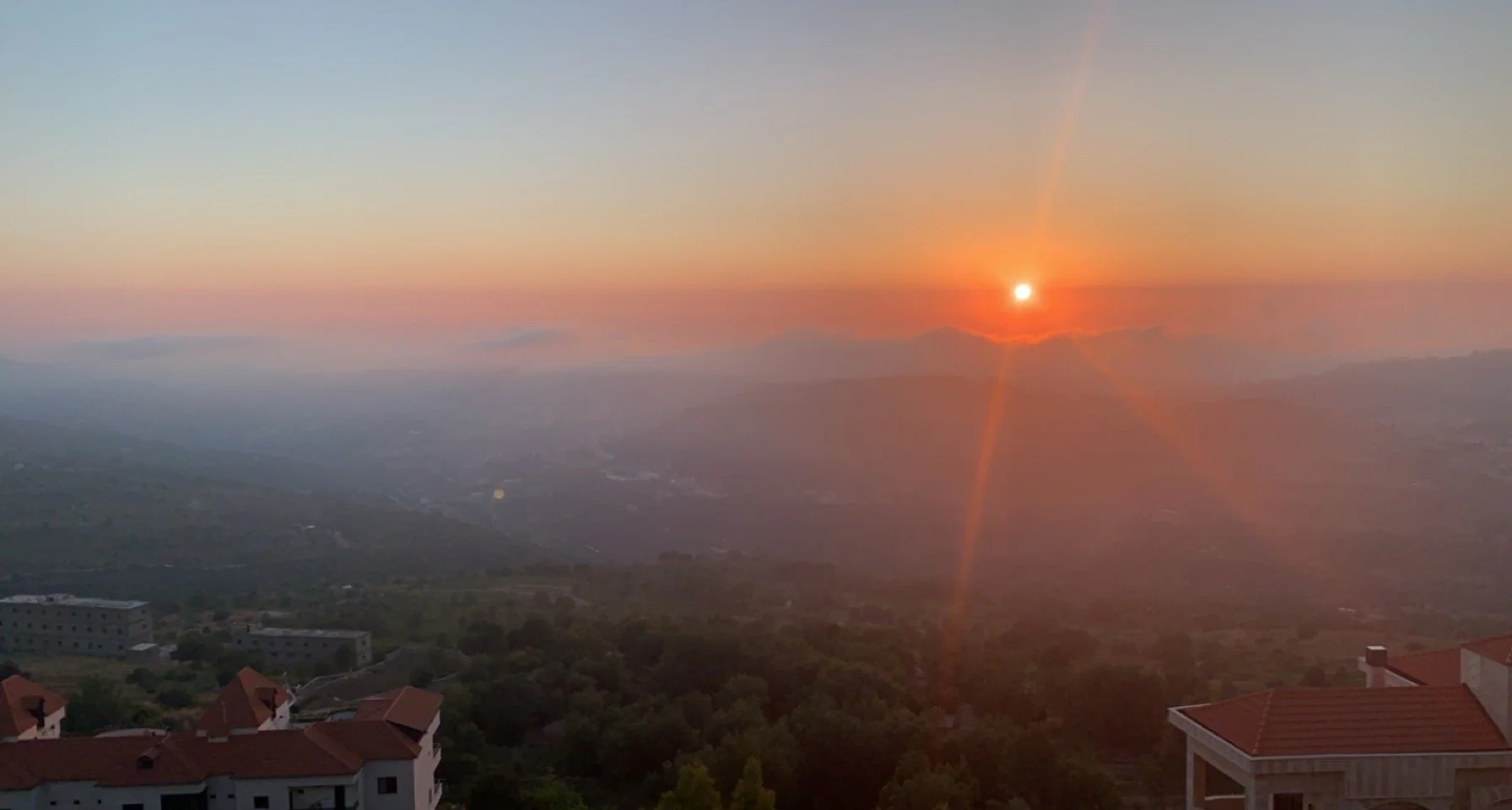 A sunset over a hilly landscape viewed from a high vantage point, with rooftops of houses visible in the foreground and a colorful pink and orange sky.
