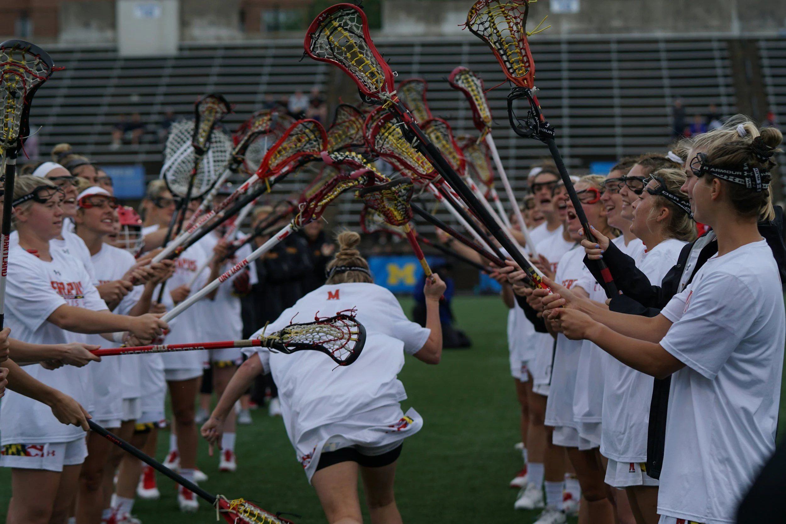 A group of female lacrosse players in white uniforms holding lacrosse sticks, gathered outdoors on a field, with some players wearing protective glasses and headgear, engaging in a team activity or celebration.