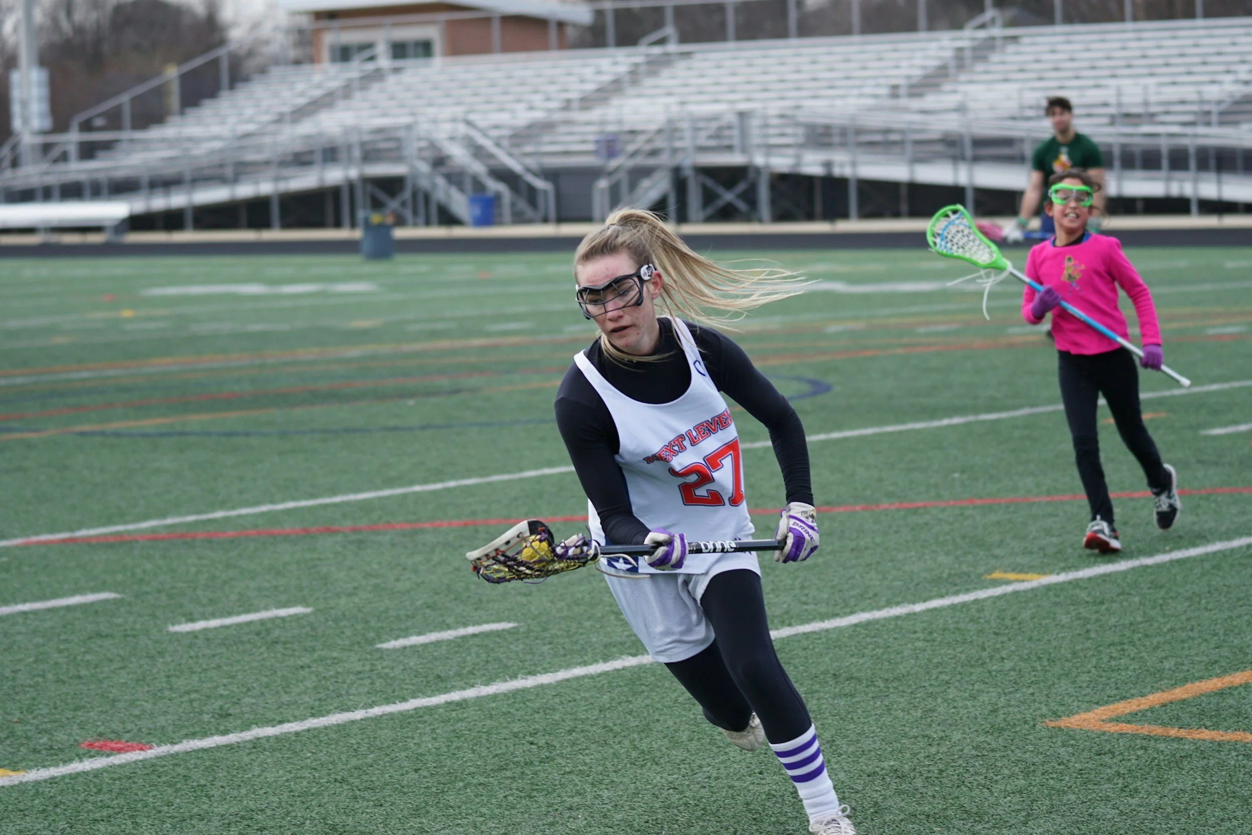 Young girls playing girls lacrosse on a sports field, one girl in a black and white uniform with a lacrosse stick, and another girl in a pink sweatshirt with protective goggles, holding a lacrosse stick, with empty bleachers in the background.