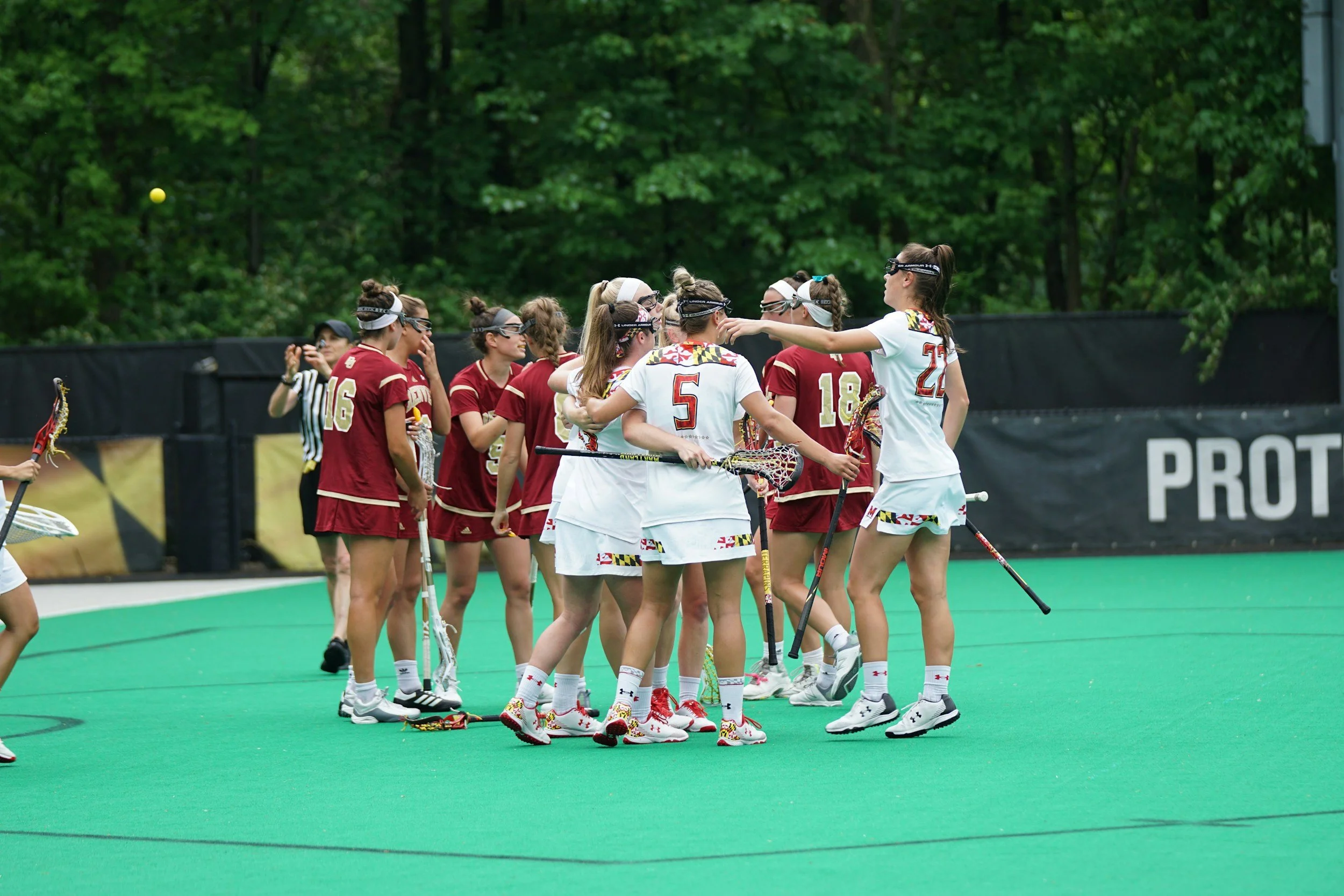 A group of female lacrosse players in red and white uniforms gather on a green field during a game, with some wearing goggles and holding lacrosse sticks, and a tennis ball in the air.