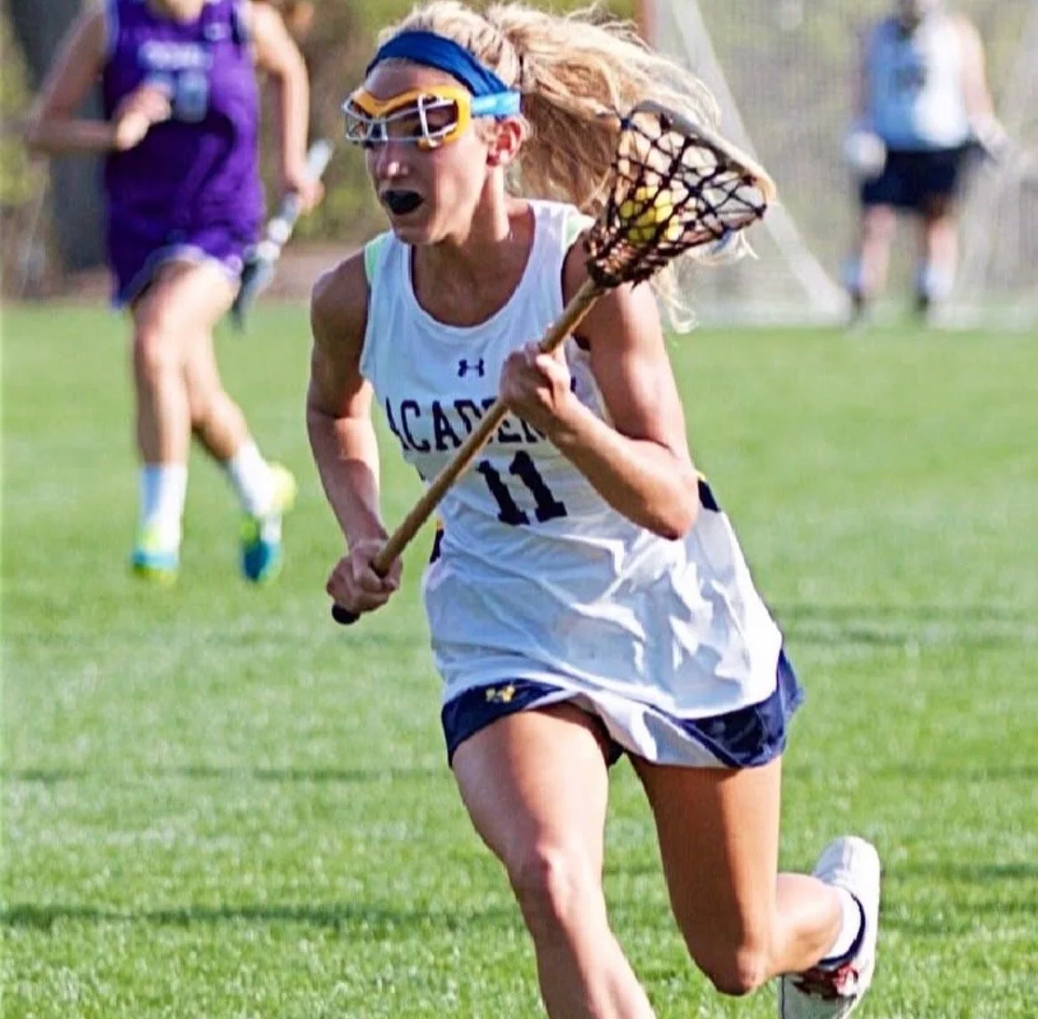 A female lacrosse player in a white and navy uniform running on a grassy field, holding a lacrosse stick, wearing protective goggles and a headband, with her hair flowing behind her.