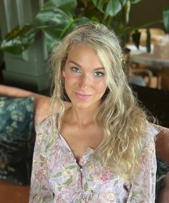 A girls lacrosse coach with long, curly blonde hair sitting on a couch, wearing a coastal blouse with pastel colors, in a lacrosse room with great lacrosse skills in the background.