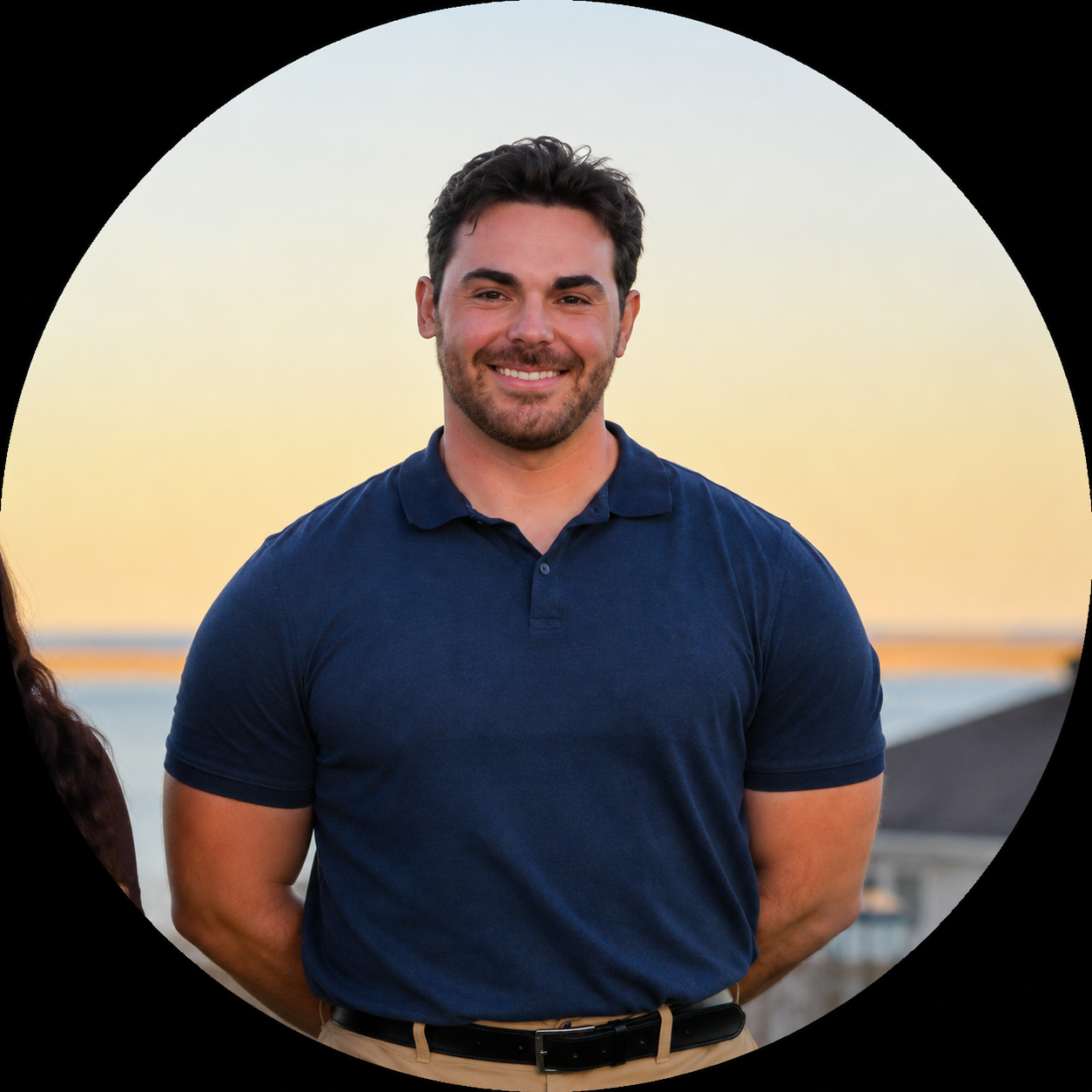A kind and caring lacrosse coach with short dark hair, beard, and mustache, wearing a navy blue polo shirt and beige pants, standing at a lacrosse banquet with athletes on Cape Cod during sunset with a blurred background of water and sky.