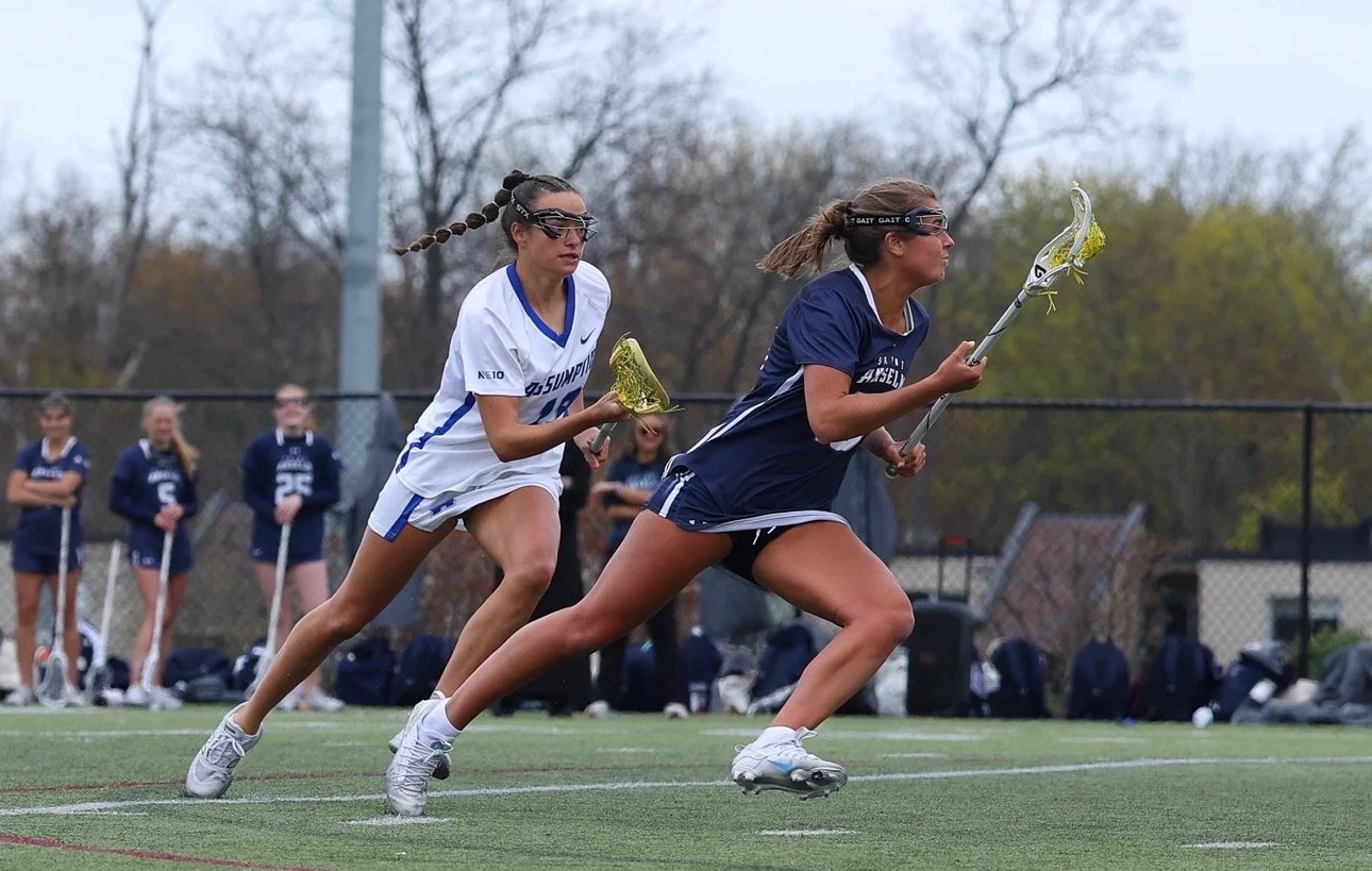 Two women playing lacrosse on a field, running with their lacrosse sticks. One in a white jersey and the other in a navy blue jersey, with other players and a chain-link fence in the background.