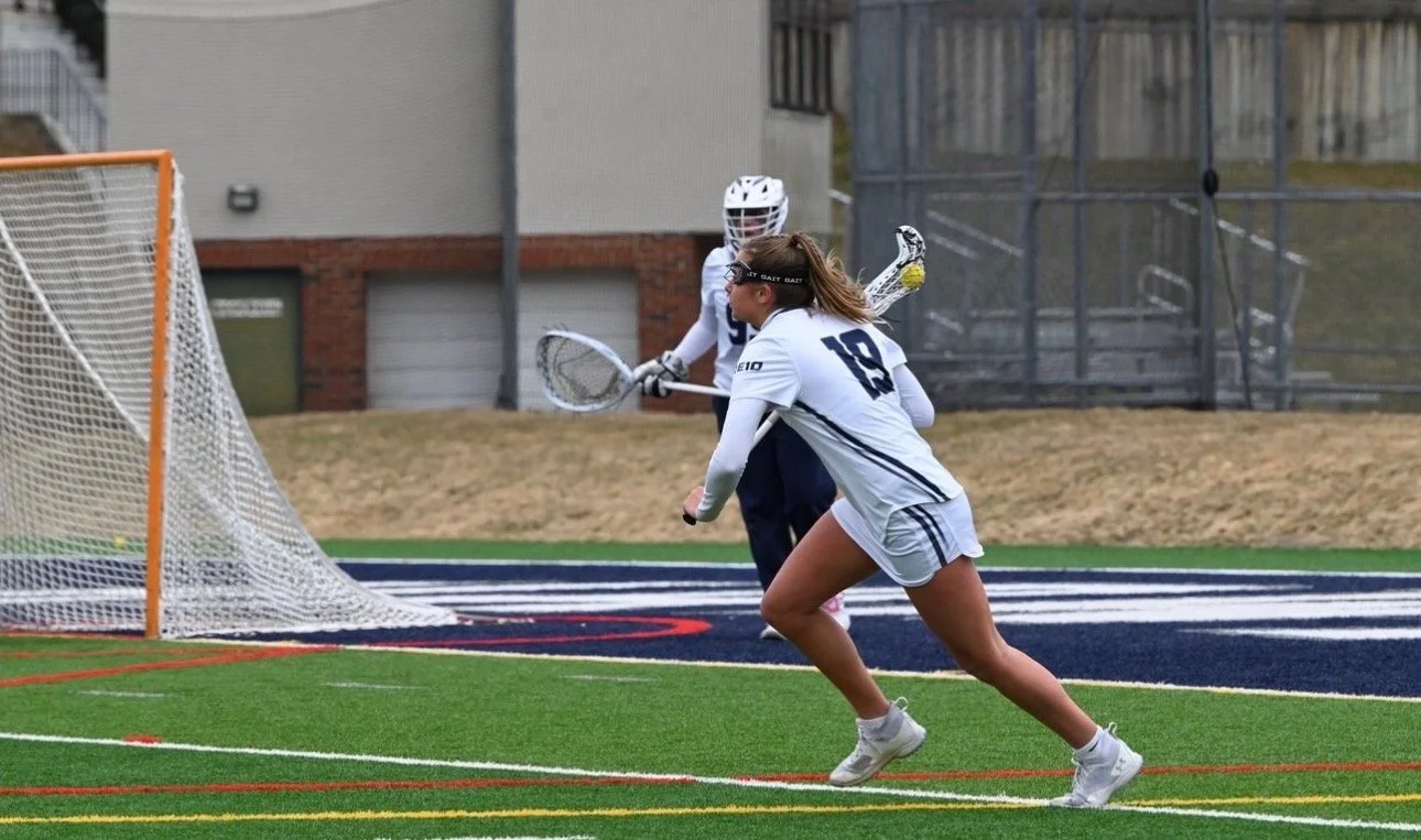 A female lacrosse player in white uniform and goggles running towards the goal with a goalie in the background on a turf field.