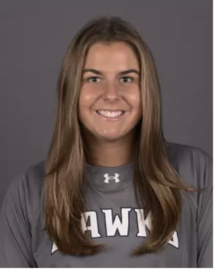 Young, kind, and caring woman lacrosse player and coach with long brown hair wearing a gray College NCAA lacrosse jersey with white text and a logo, smiling against a gray background.