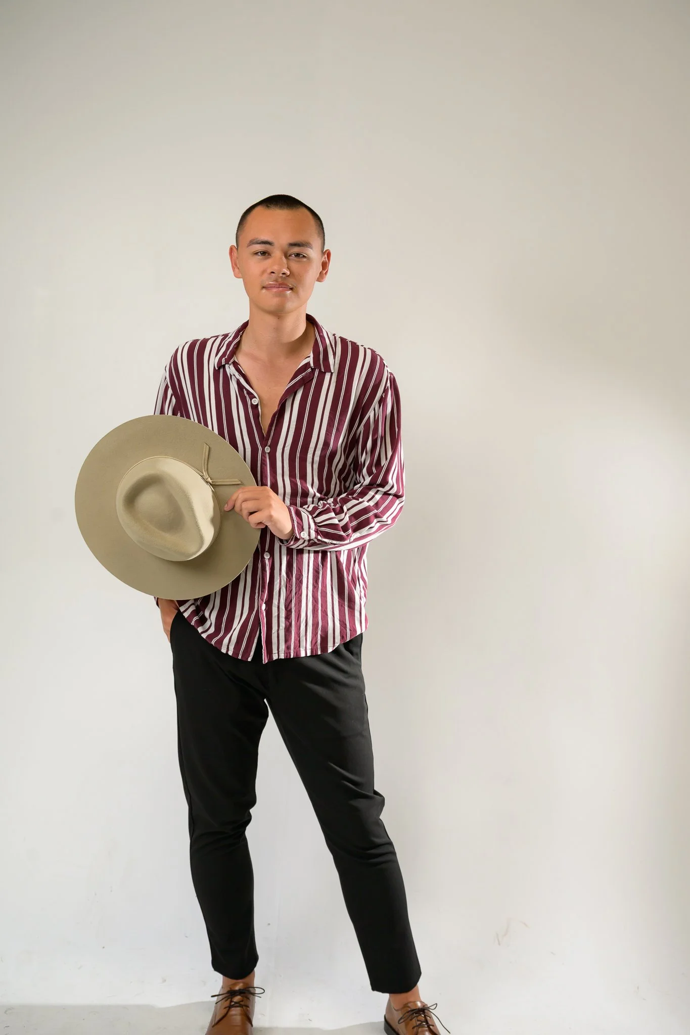 A young man with a mohawk hairstyle, wearing a striped maroon and white shirt, black pants, and tan shoes, holding a beige hat, standing against a plain white wall.