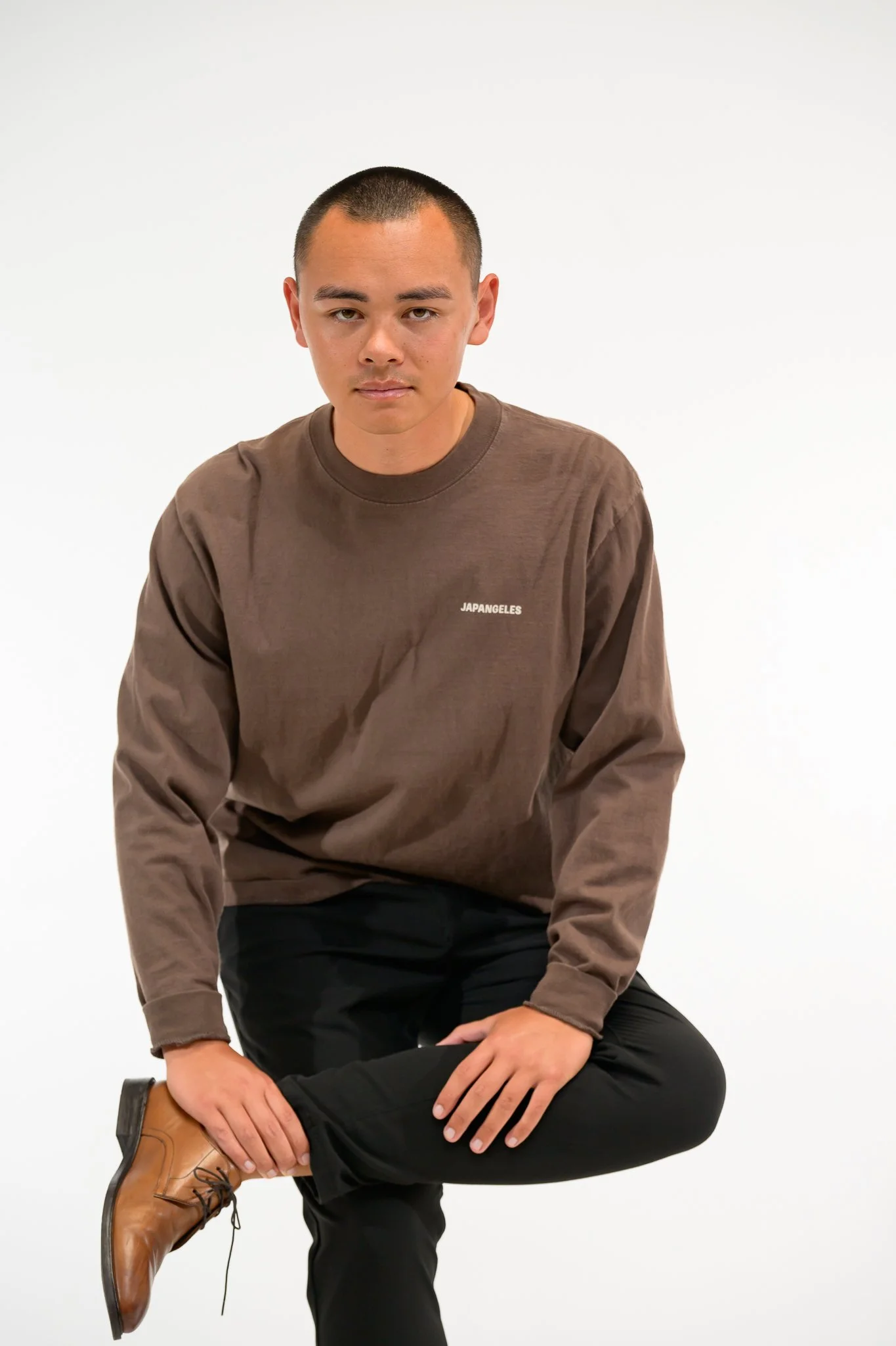 A young man with short hair and light skin sitting on a stool against a plain white background. He is wearing a brown sweatshirt with a small white logo on the chest, black pants, and tan boots. He is looking directly at the camera with a neutral expression, one hand resting on his knee, and the other on his ankle.