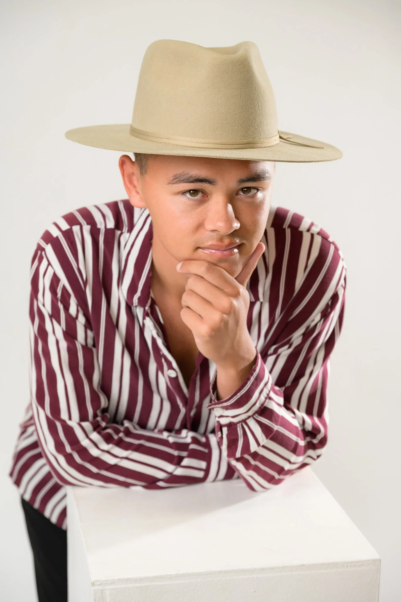A man wearing a beige fedora hat with a ribbon around it, a maroon and white striped button-up shirt, leaning on a white surface, looking directly at the camera with his chin resting on his hand.