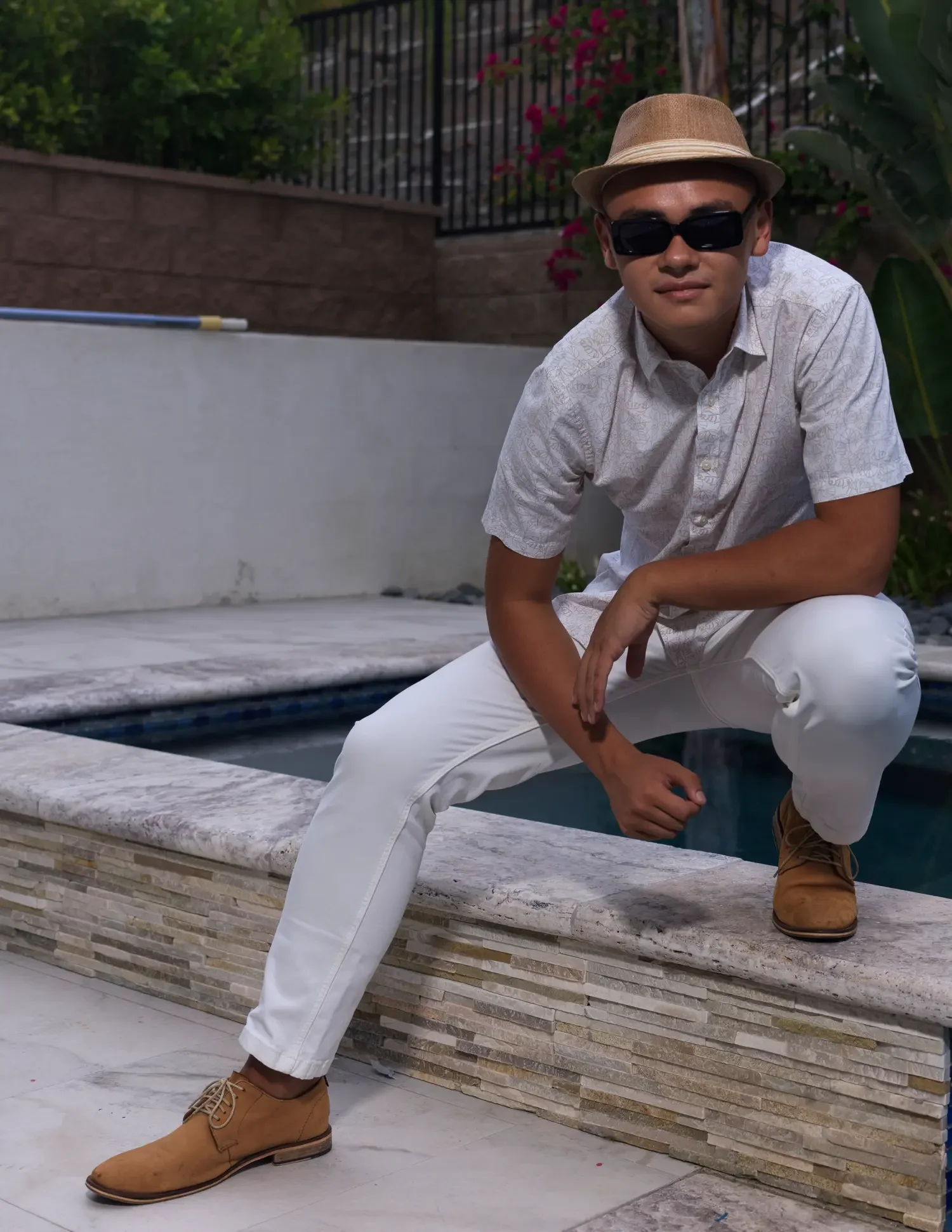 A young man wearing a beige fedora, black sunglasses, a white patterned short-sleeve shirt, white pants, and tan work boots crouching by a poolside.