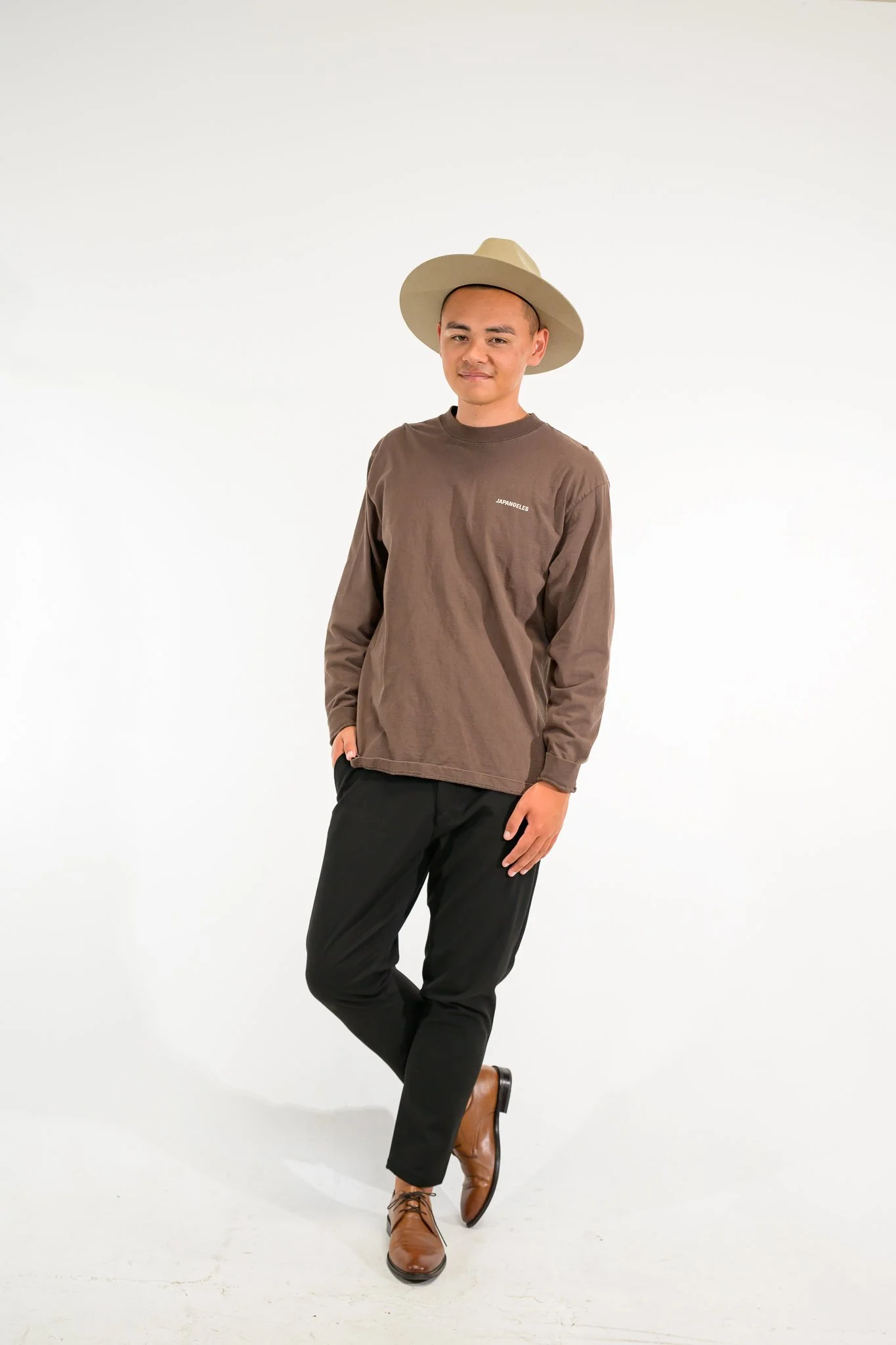 A young man in a brown long-sleeve shirt, black pants, and brown boots standing against a plain white background. He is wearing a wide beige hat and has one hand in his pocket with a slight smile.