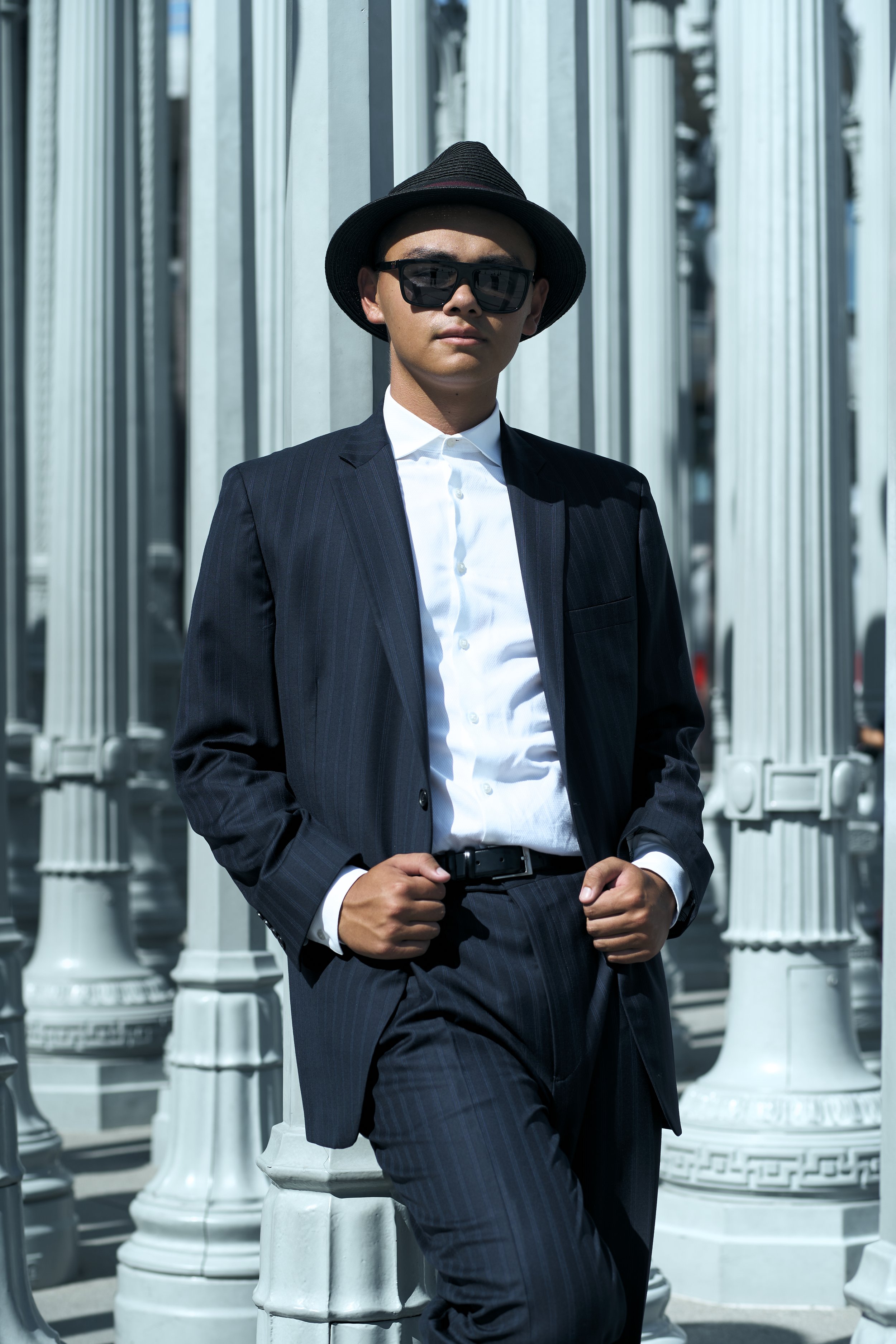 Young man in a dark pinstripe suit, white shirt, sunglasses, and fedora hat leaning against white decorative columns outdoors.