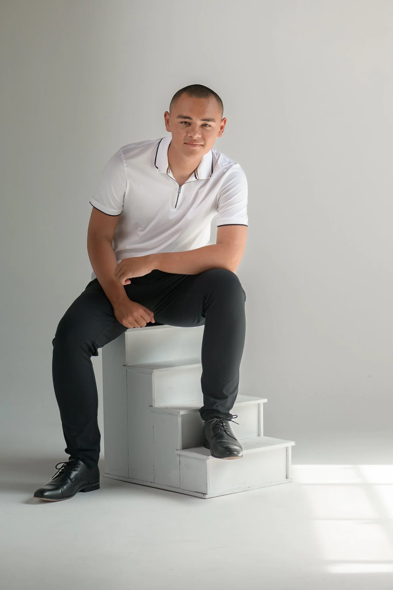 A young man with short hair, wearing a white polo shirt, black pants, and black shoes, sitting on a white set of stairs against a plain gray background.