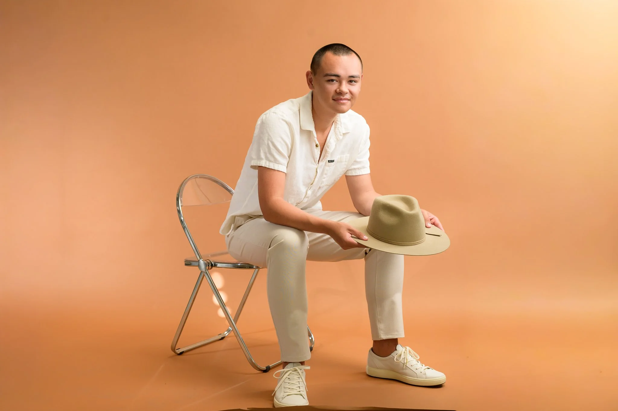 Young man with short hair sitting on a clear plastic and metal chair against a peach-colored background, holding a beige wide-brimmed hat, dressed in a white short-sleeved shirt, beige pants, and white sneakers.