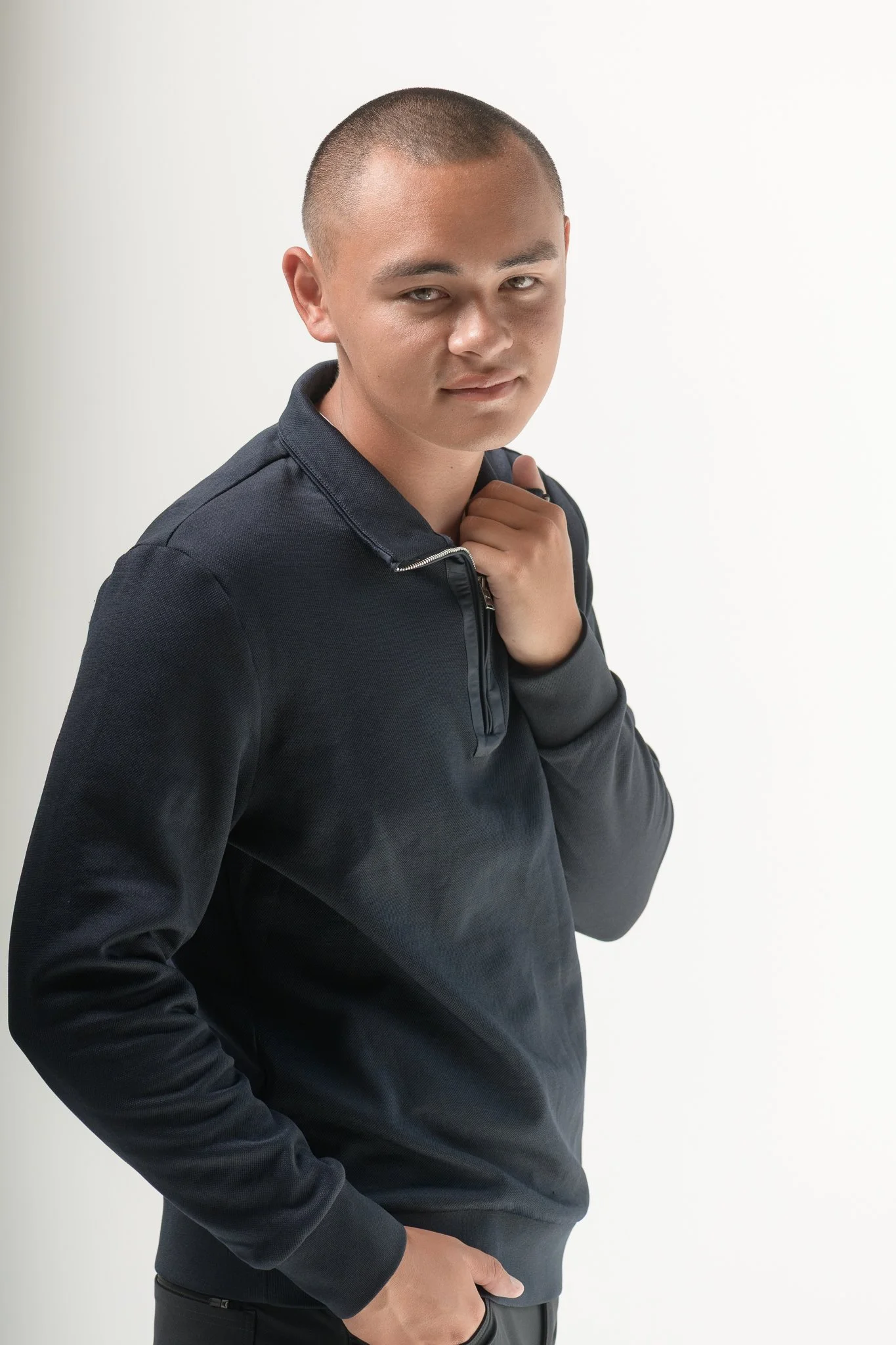 A young man with short hair and a confident expression, wearing a dark zip-up jacket and black pants, standing against a plain white background.