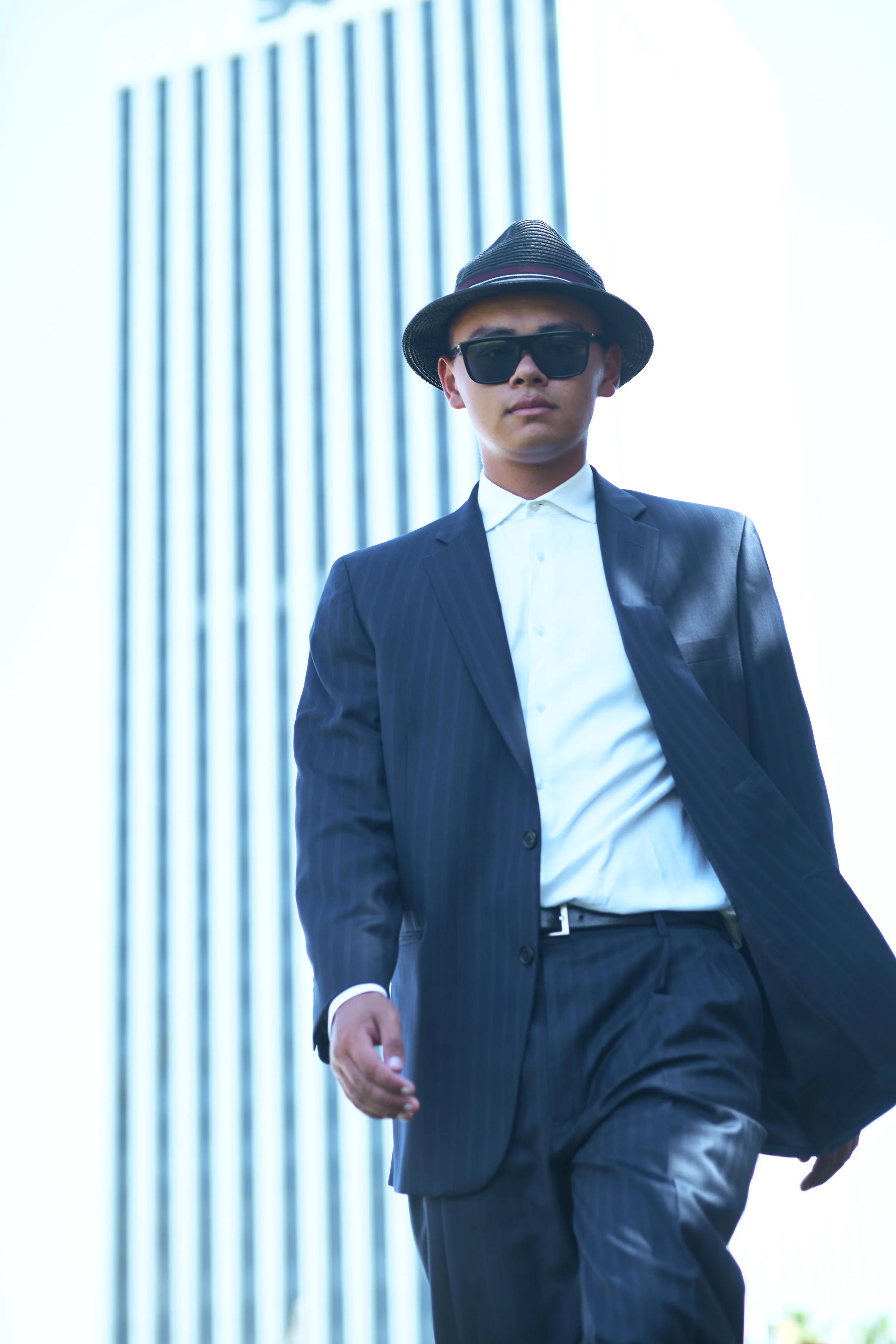 A man in a dark pinstripe suit, white shirt, and black sunglasses, wearing a fedora hat, walking outdoors in front of a modern building with vertical striped windows.