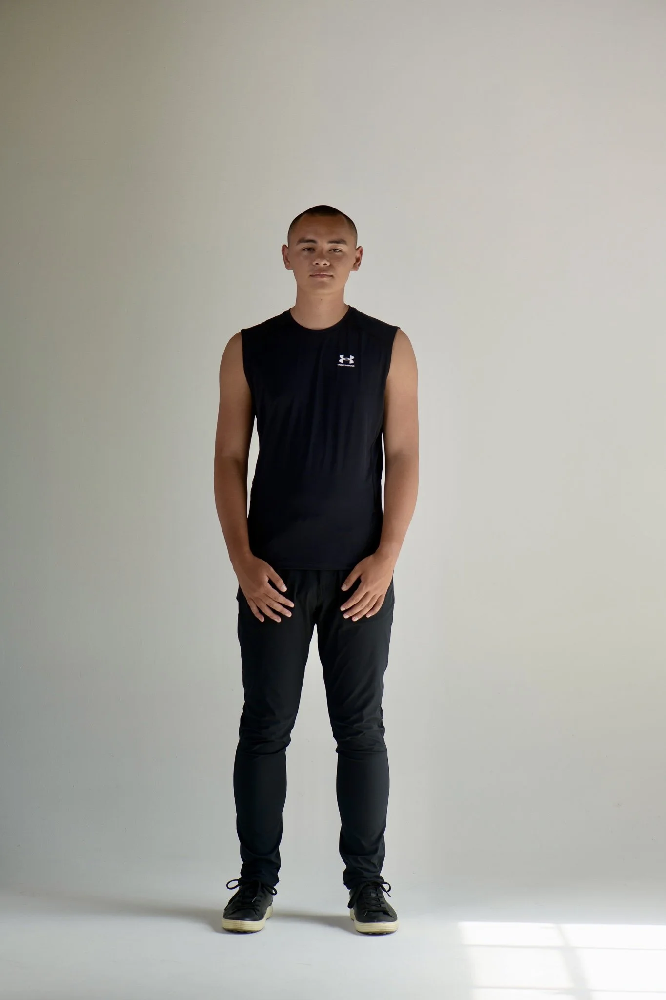 A young man standing in front of a plain white wall, wearing a black sleeveless athletic shirt, black pants, and sneakers, with short hair and a neutral expression.