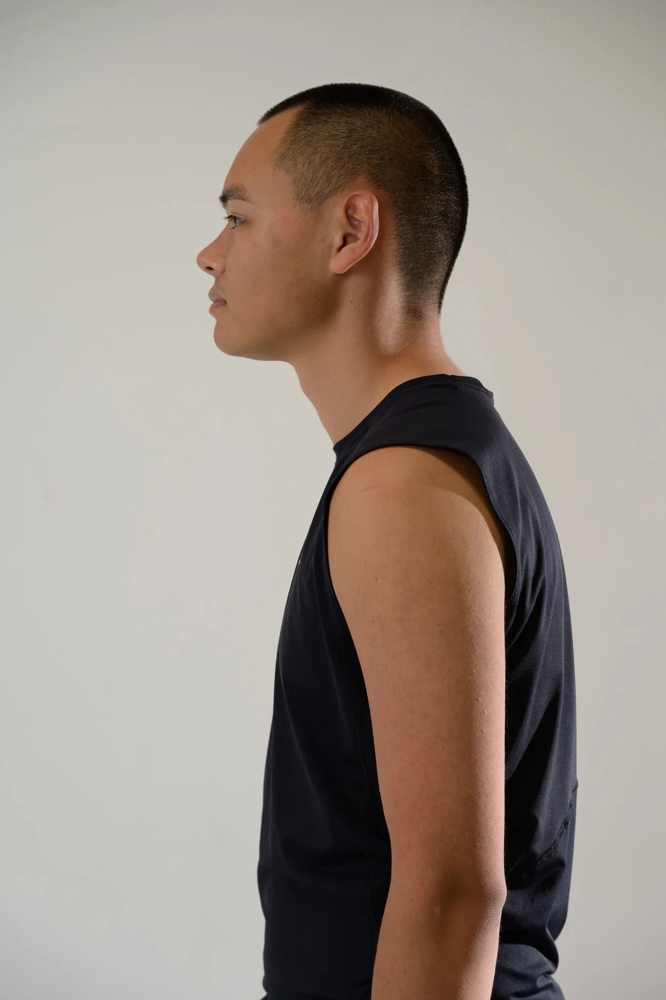 Side view of a young man with a sport haircut wearing a black sleeveless shirt against a plain light-colored background.