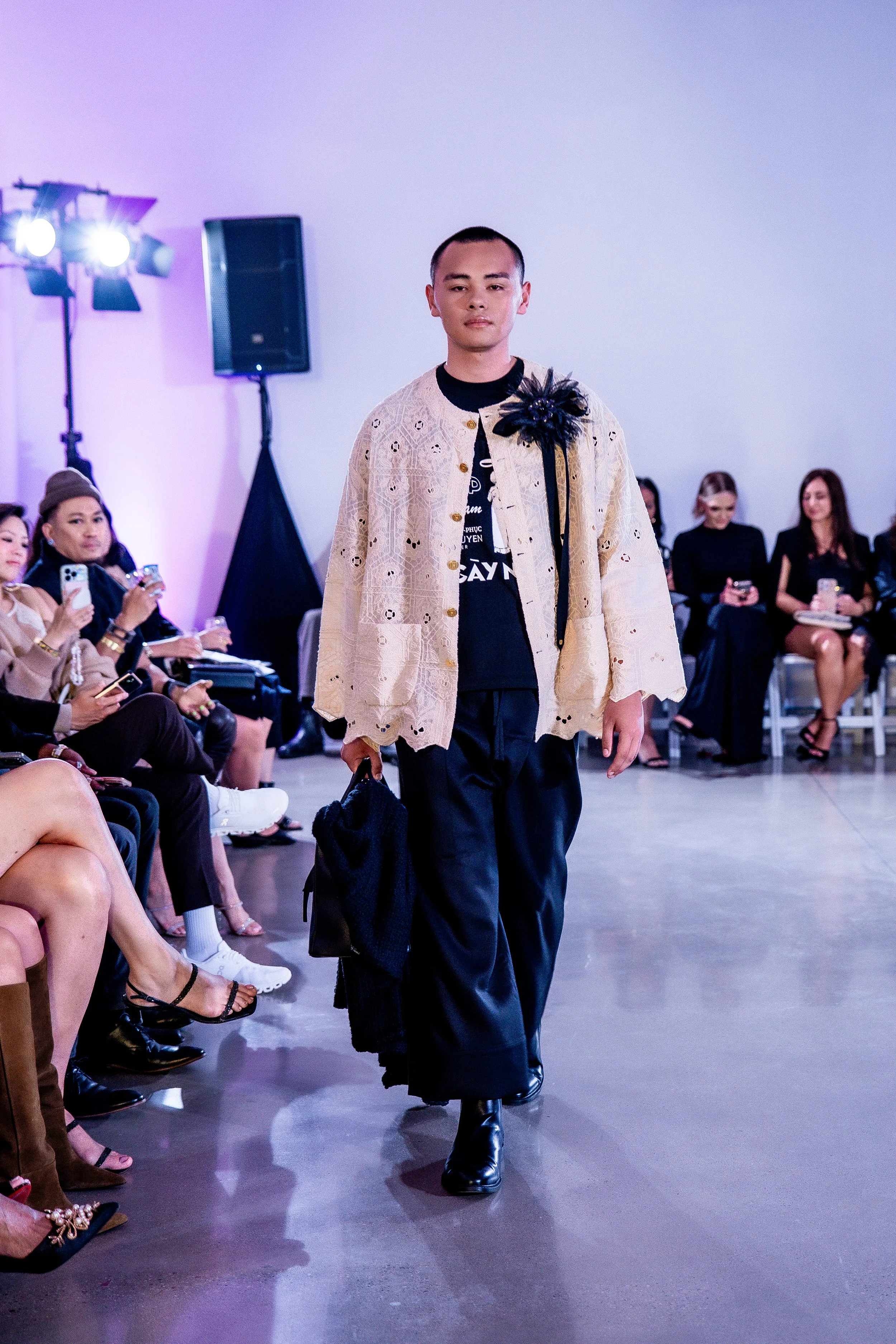 A male model walks down the runway during a fashion show, wearing a cream-colored coat with eyelet details and black pants, in front of an audience of seated spectators.