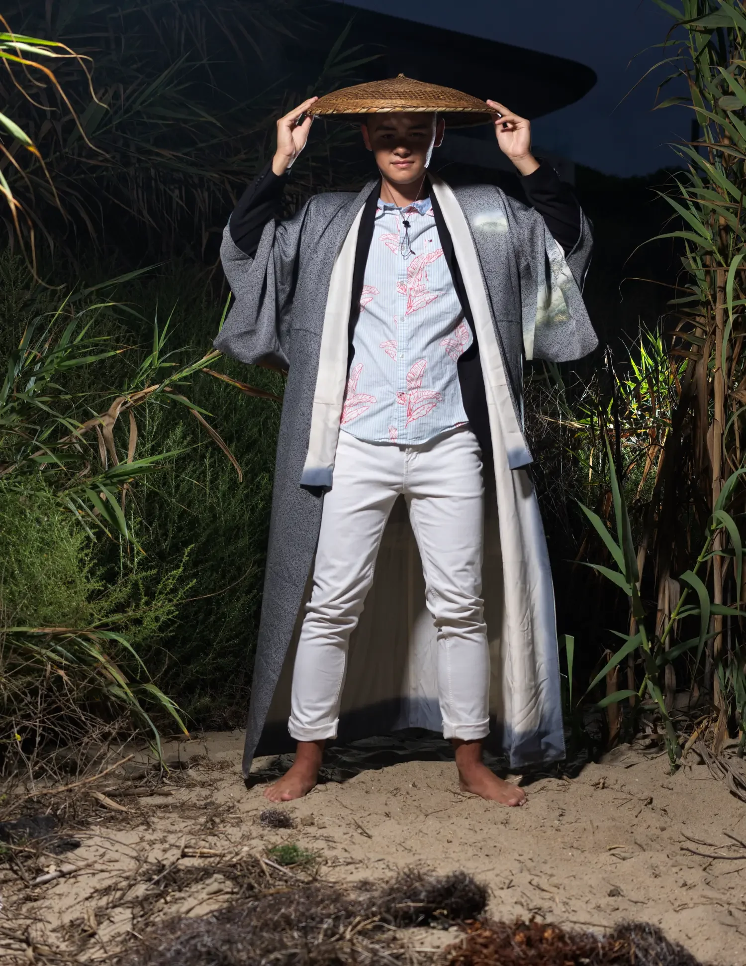 Person standing barefoot on sand, wearing a patterned shirt, white pants, a gray coat, and holding a conical hat on their head, surrounded by tall grass and plants at night.