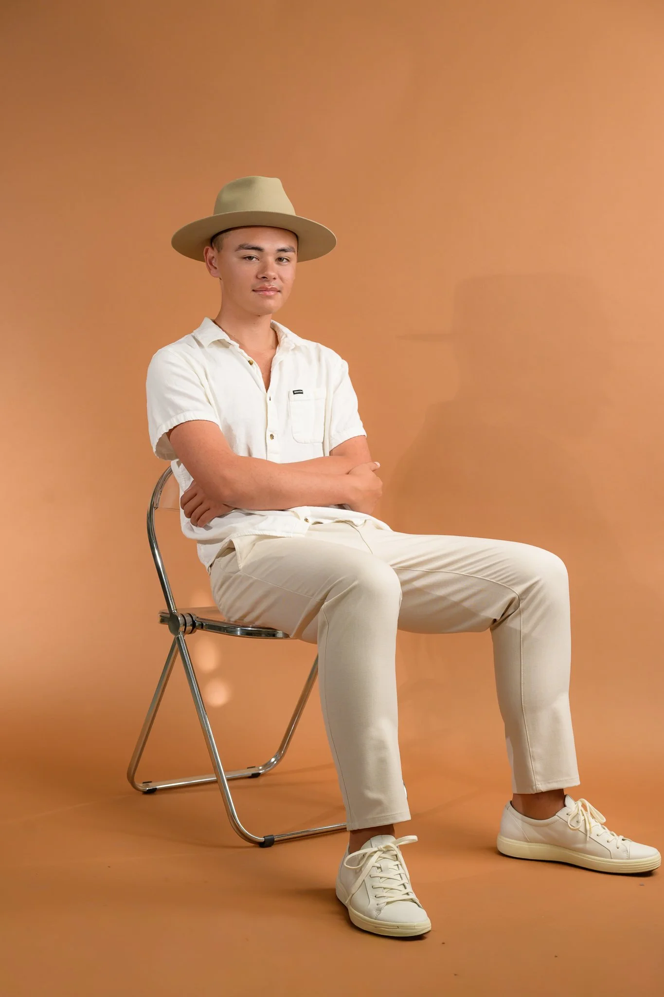 A young man sitting on a folding chair with a beige background. He is wearing a beige wide-brimmed hat, a white short-sleeve button-up shirt, cream-colored pants, and white sneakers. His arms are crossed, and he has a neutral expression.