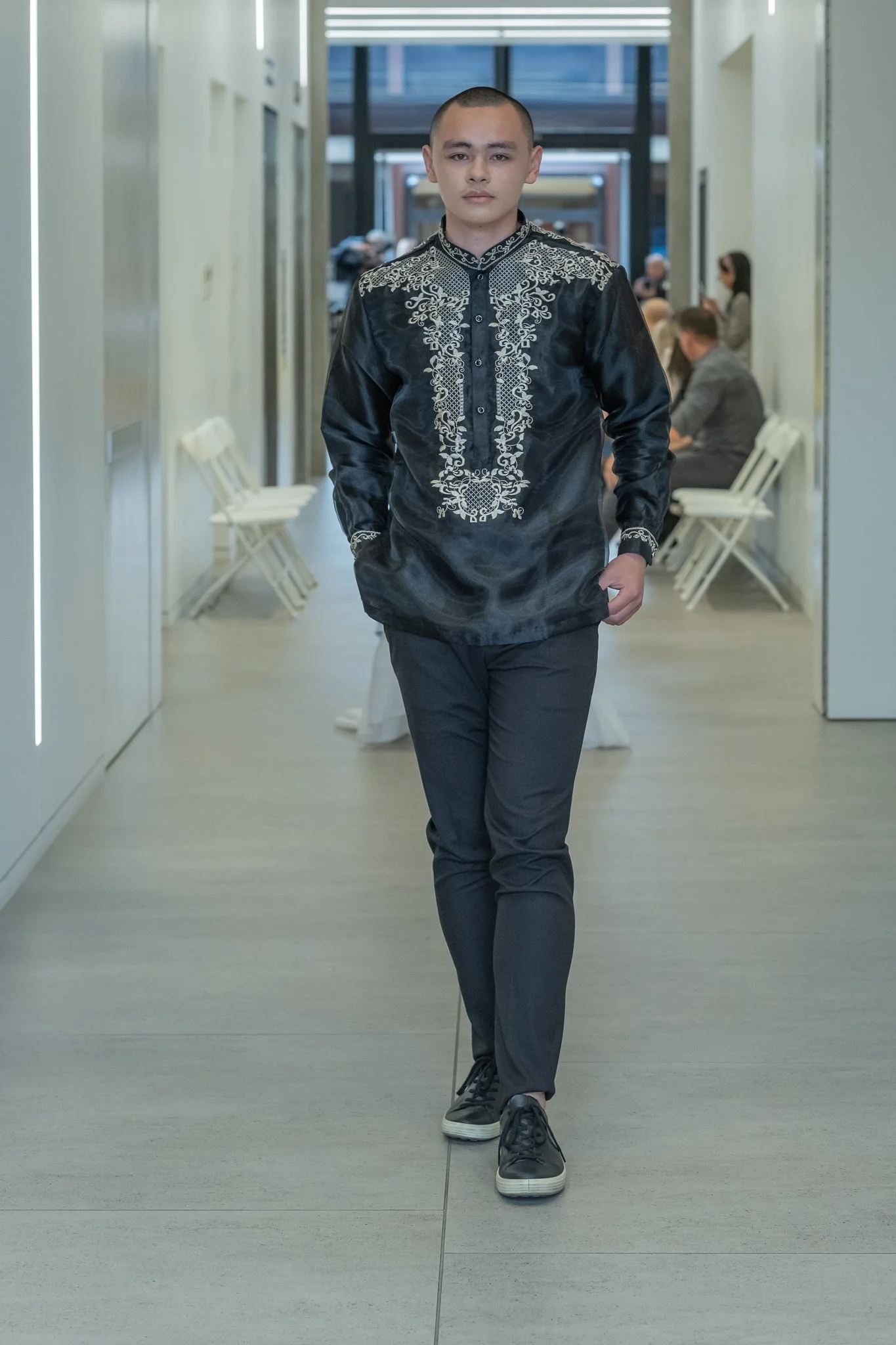 A young man walking through a modern building corridor with large glass windows and seated people in the background.