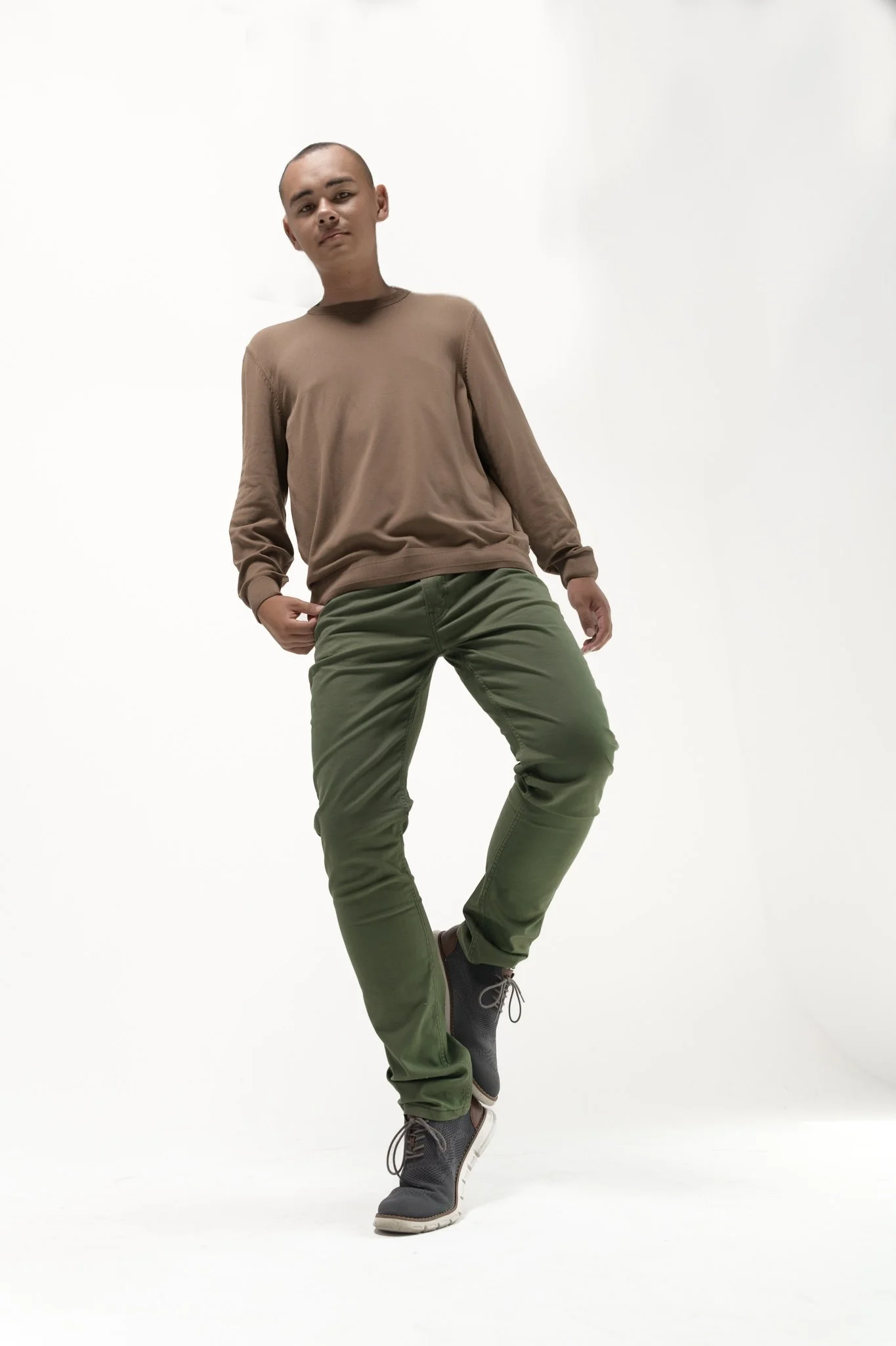 A young person with a shaved head poses confidently in a studio with a plain white background. They are wearing a brown long-sleeve shirt, green pants, and gray shoes.