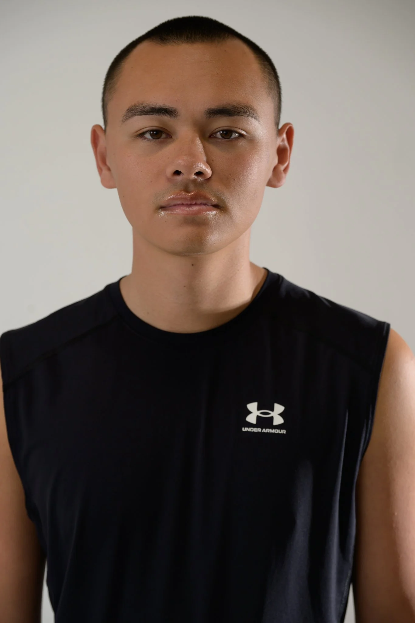 Close-up of a young man with short hair wearing a black Under Armour sleeveless athletic shirt, standing against a plain light-colored background.