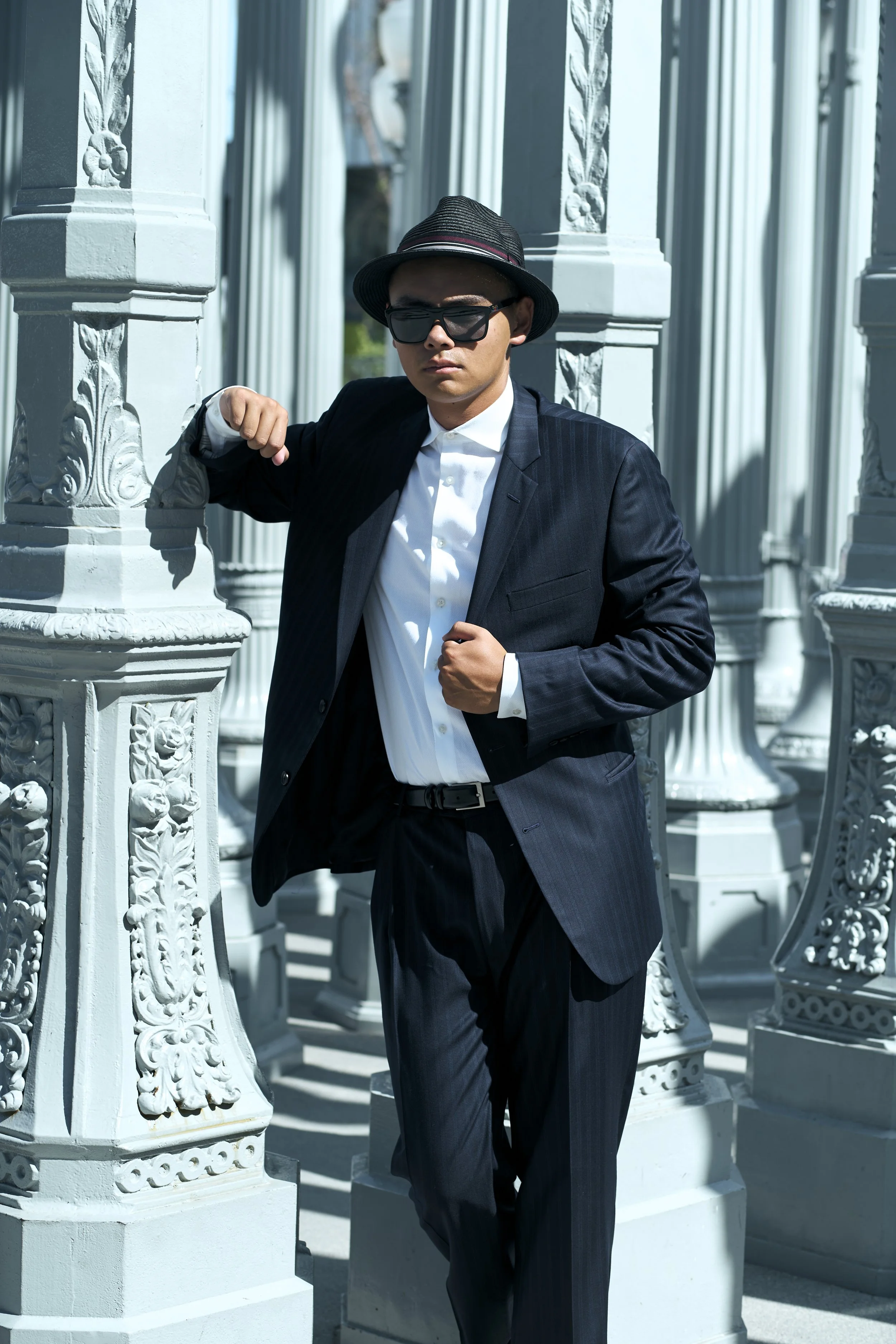 A young man dressed in a black pinstripe suit, white shirt, and black hat stands outdoors among ornate white columns. He wears sunglasses and leans casually against one of the columns with his right arm, looking confidently at the camera.