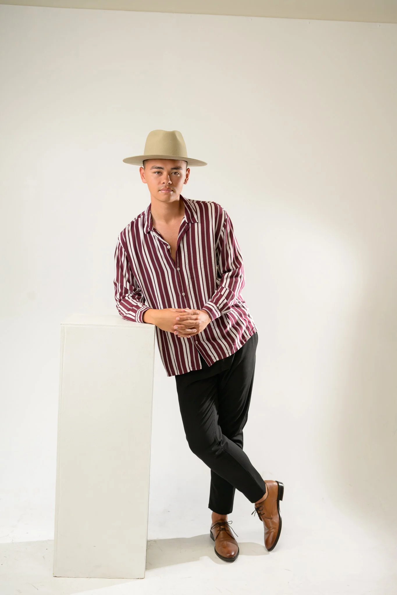 A person leaning against a white rectangular block, wearing a beige wide-brimmed hat, a red and white striped button-up shirt, black pants, and brown lace-up boots, standing against a plain light-colored background.