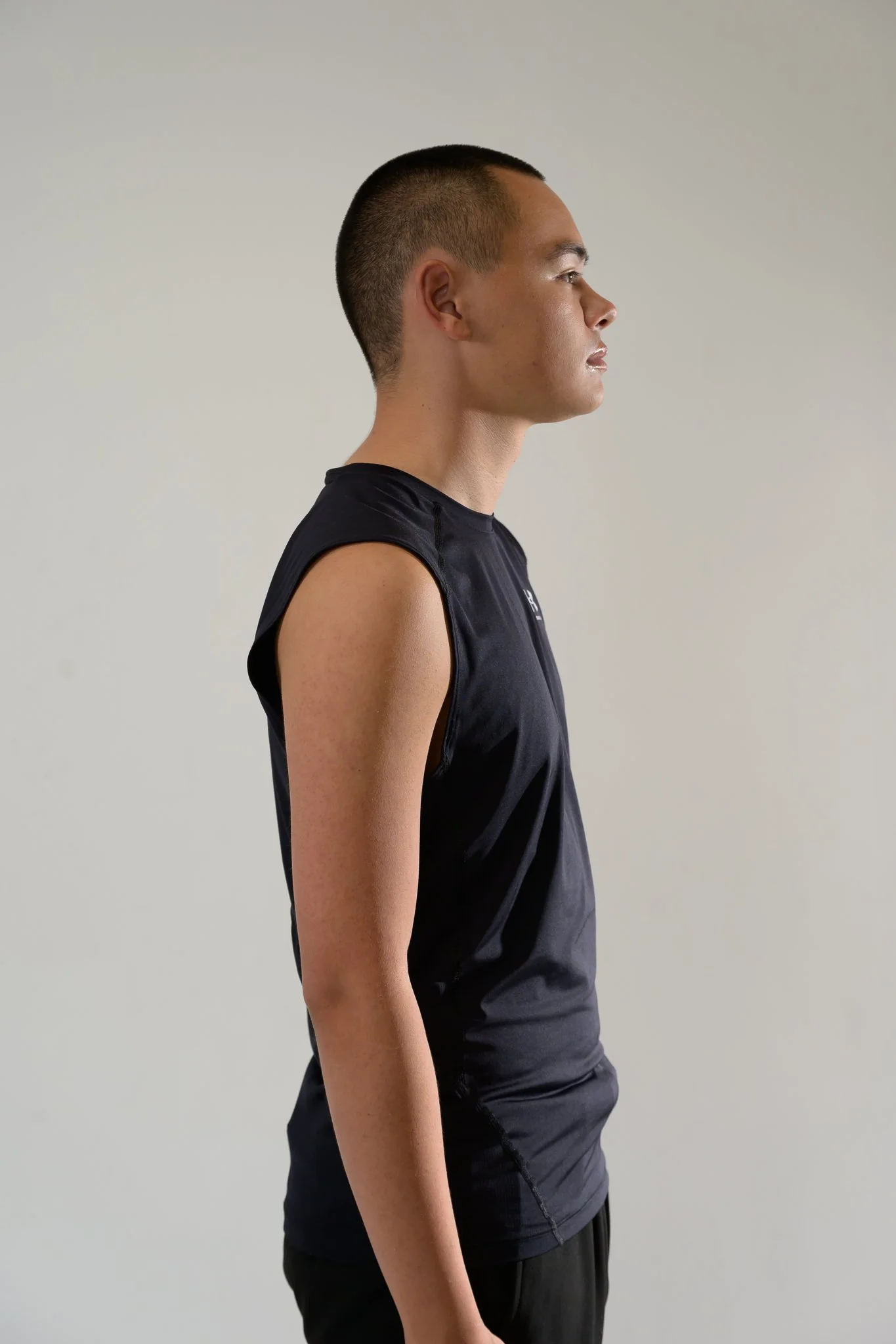 Side view of a young man wearing a black sleeveless athletic shirt and black shorts, standing against a plain white background.