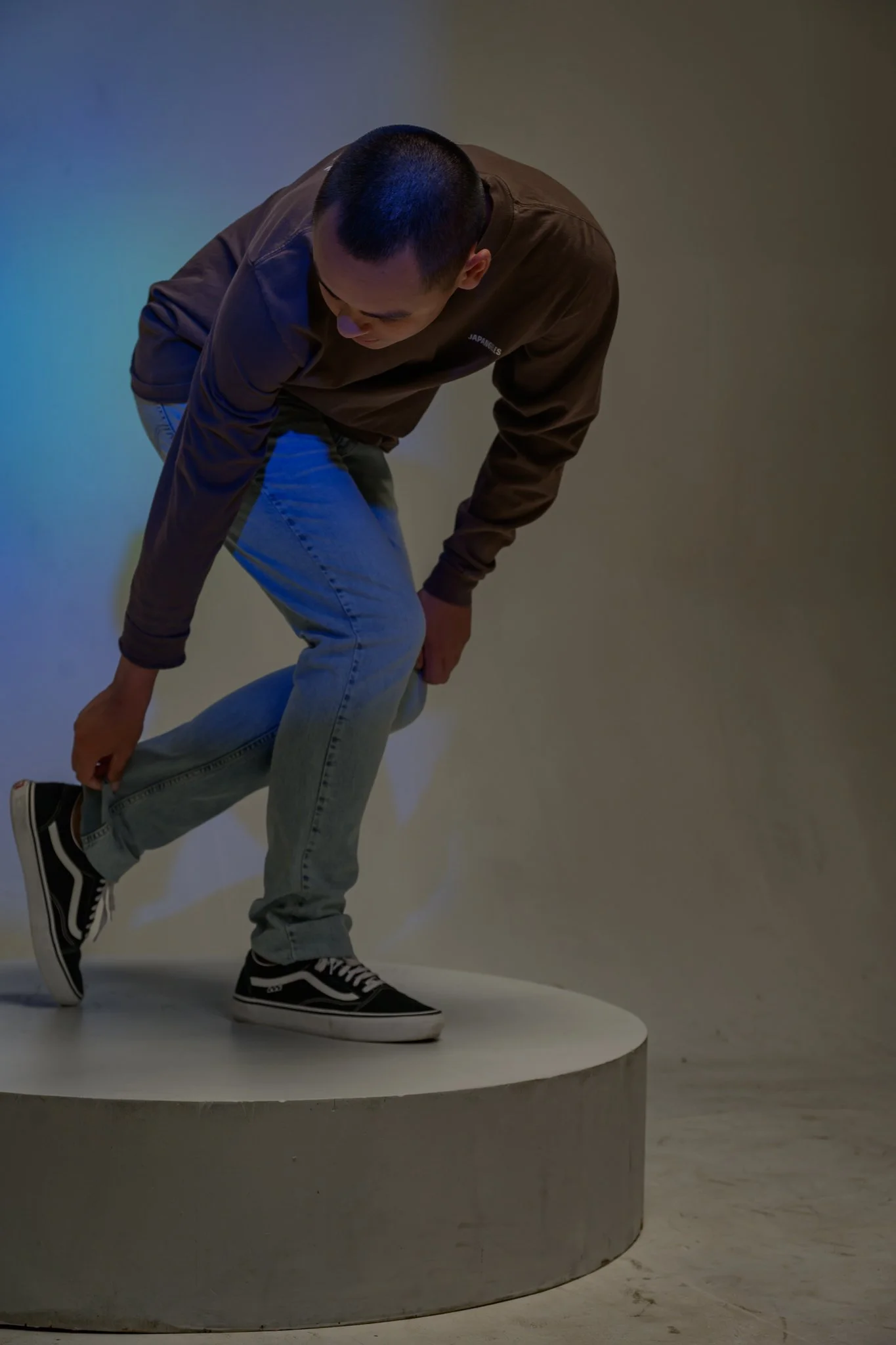 A person dressed in casual clothes, wearing Vans sneakers, bending down on a circular platform in a minimalist setting with plain walls.