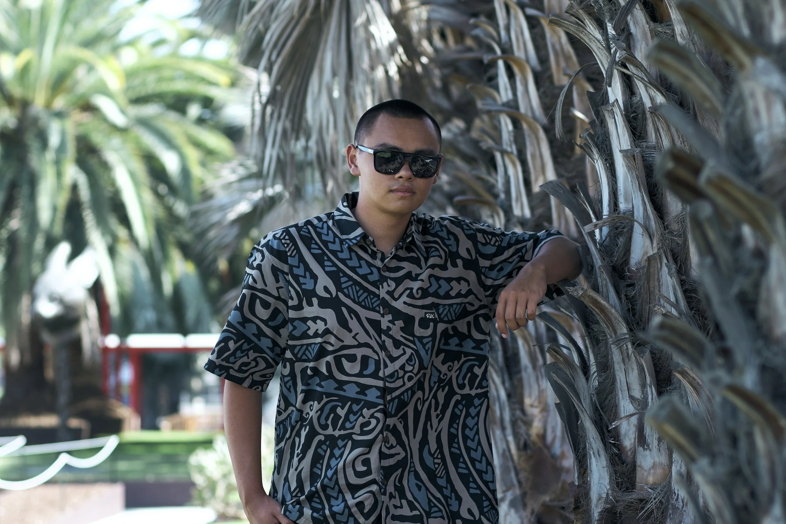 A young man wearing sunglasses and a patterned shirt standing beside a tree with large, dry leaves.