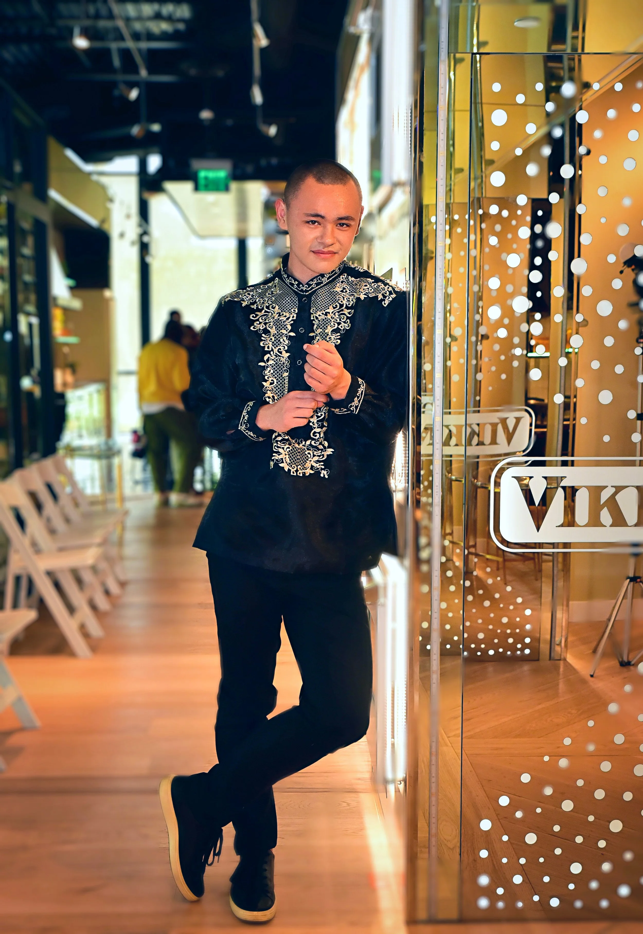 A young man with a shaved head wearing a black embroidered shirt, black pants, and black sneakers with white soles, leaning against a glass wall decorated with white polka dots inside a modern retail store.