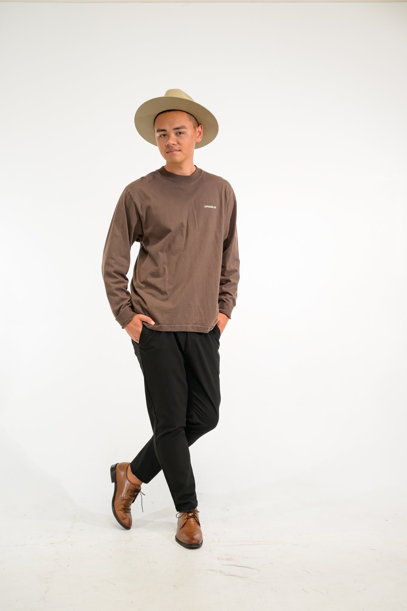 A young man wearing a wide-brimmed hat, brown long-sleeve shirt, black pants, and brown shoes standing against a plain white background. He has his hands in his pockets and is looking directly at the camera.