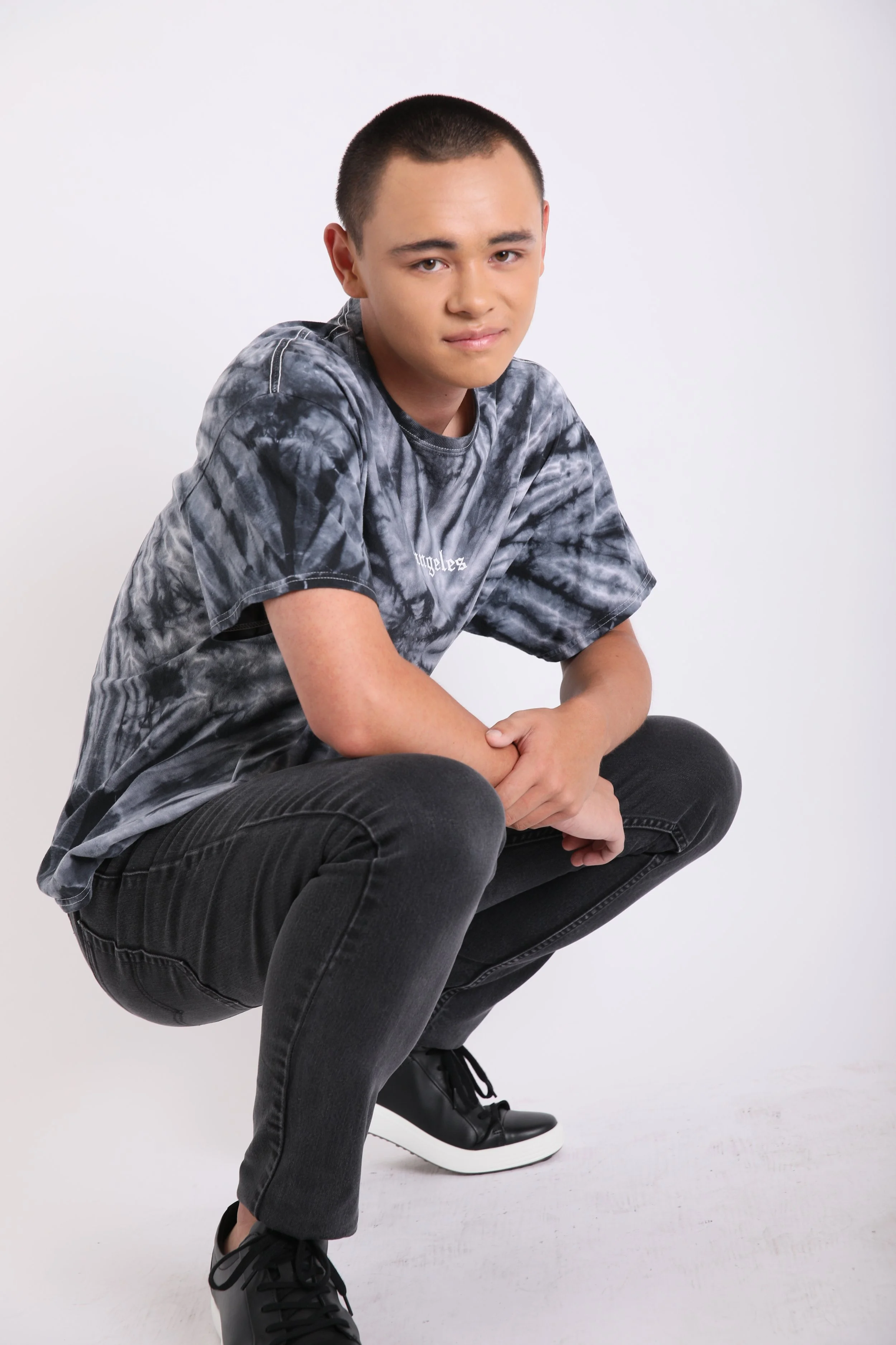 A young man with short hair and light skin, wearing a gray tie-dye T-shirt, black jeans, and black sneakers, is squatting against a plain white background.