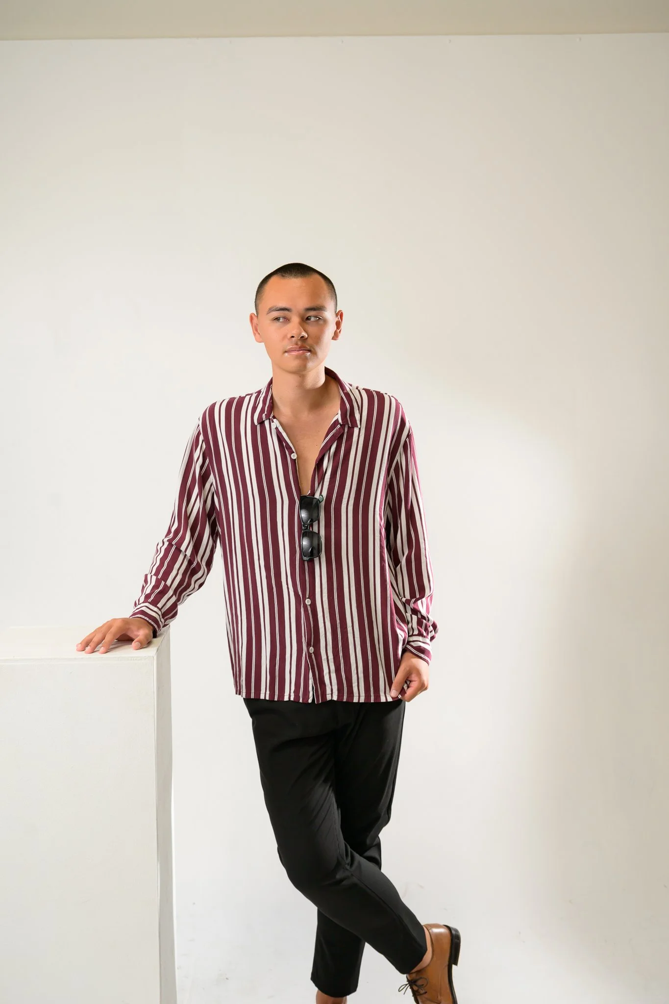 A young person with a buzz cut hairstyle, wearing a striped long sleeve shirt and black pants, standing against a plain white background, with a white pedestal nearby. They are leaning on the pedestal with one hand, have sunglasses hanging from their shirt, and are wearing brown shoes.