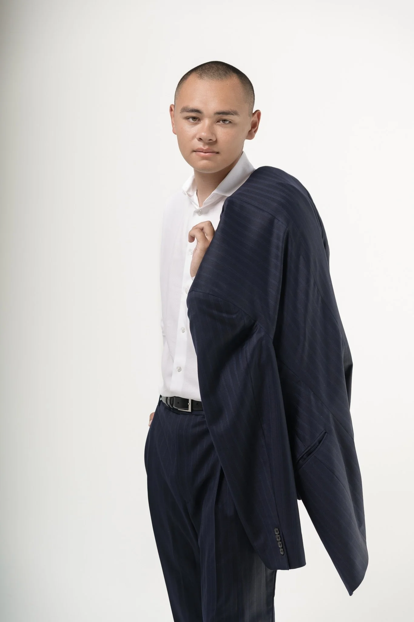 A young man with short hair wearing a white dress shirt and a dark blue suit jacket and matching pants, holding the suit jacket over his shoulder, standing against a plain white background.