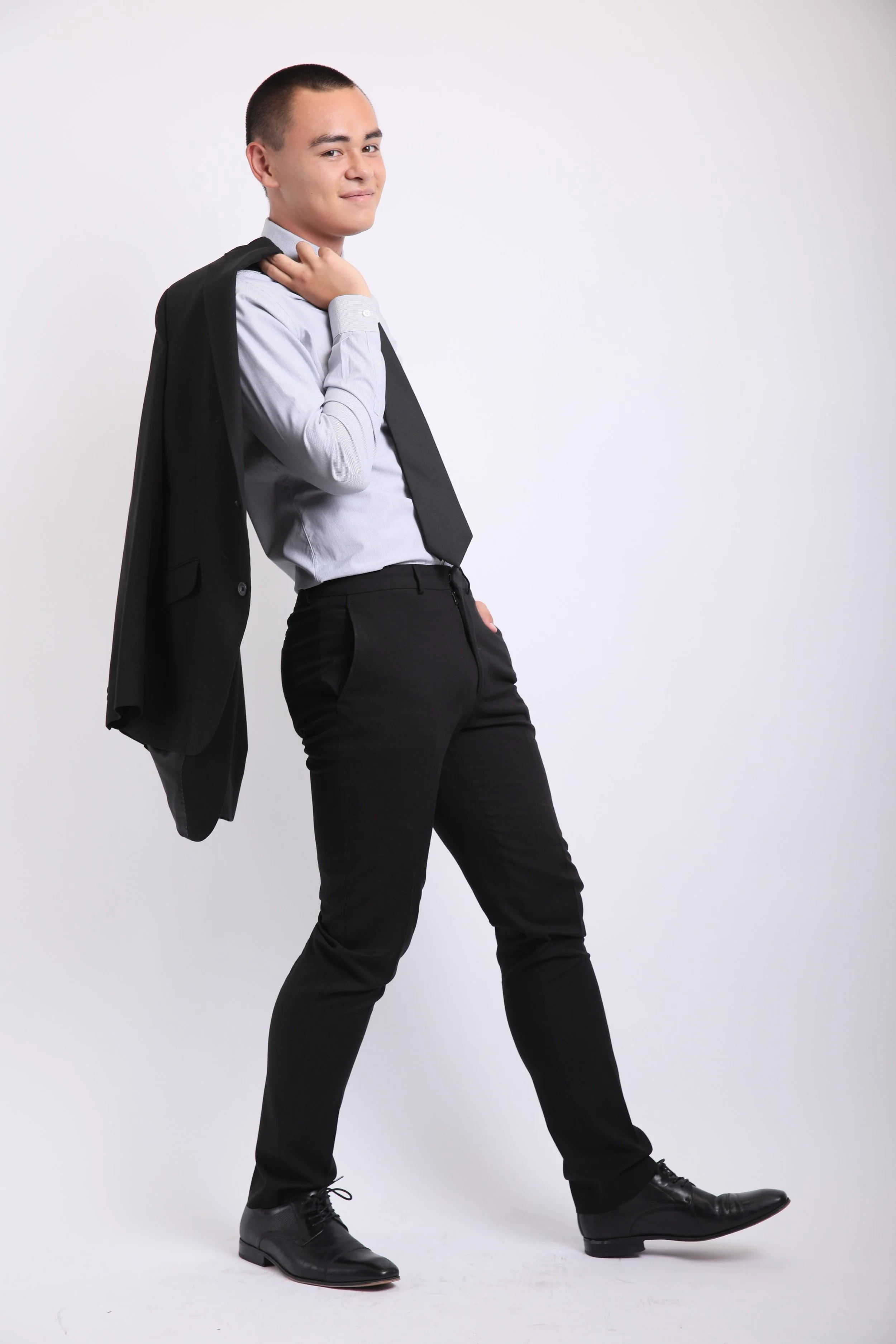 A young man standing against a plain white background, dressed in a light gray shirt, black pants, and black shoes, holding a black blazer over his shoulder with a confident expression.