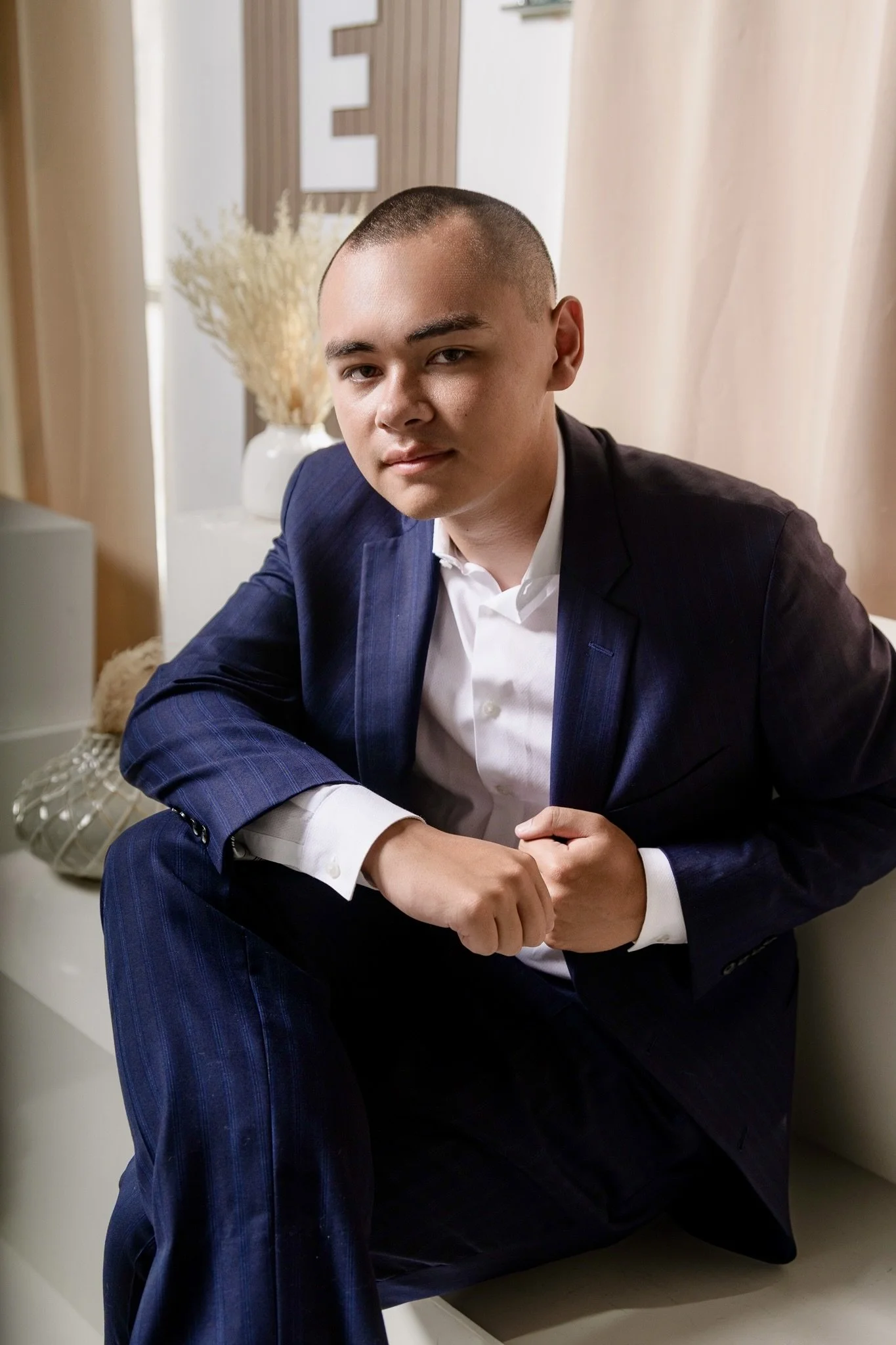 A young man in a navy blue suit and white shirt sitting on the floor, leaning against a cream-colored surface in a room with beige curtains and a decorative vase with dried plants in the background.