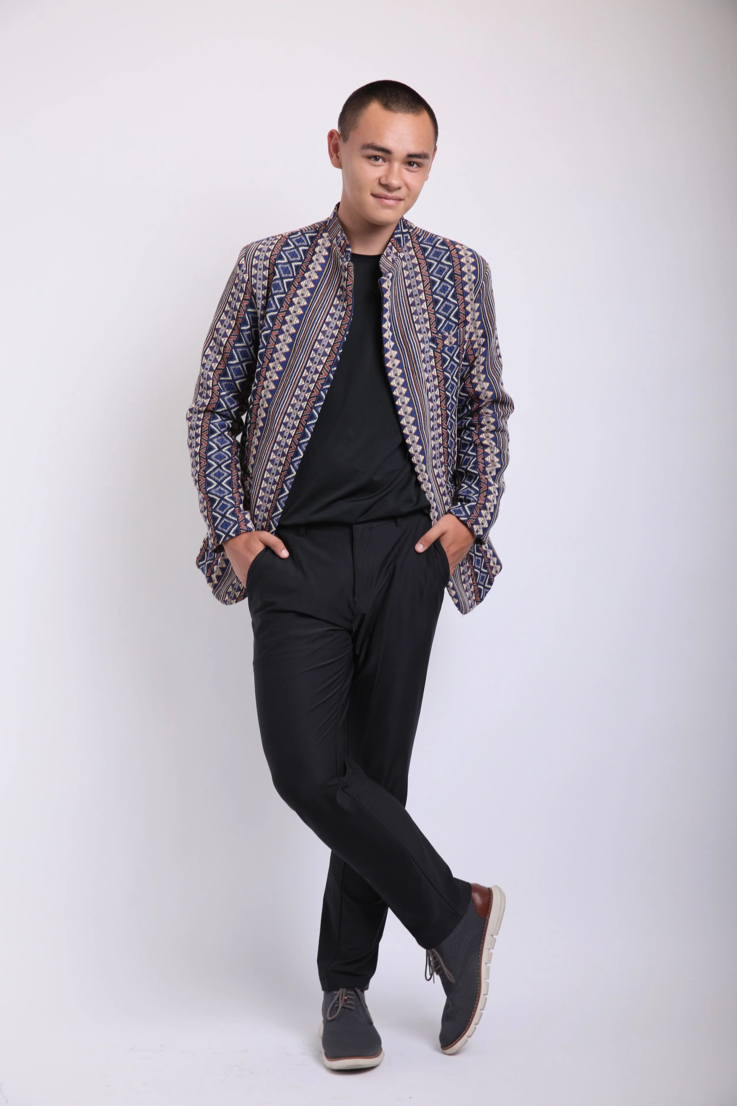 A young man with short hair wearing a patterned jacket, black shirt, black pants, and gray sneakers posing against a plain white background.